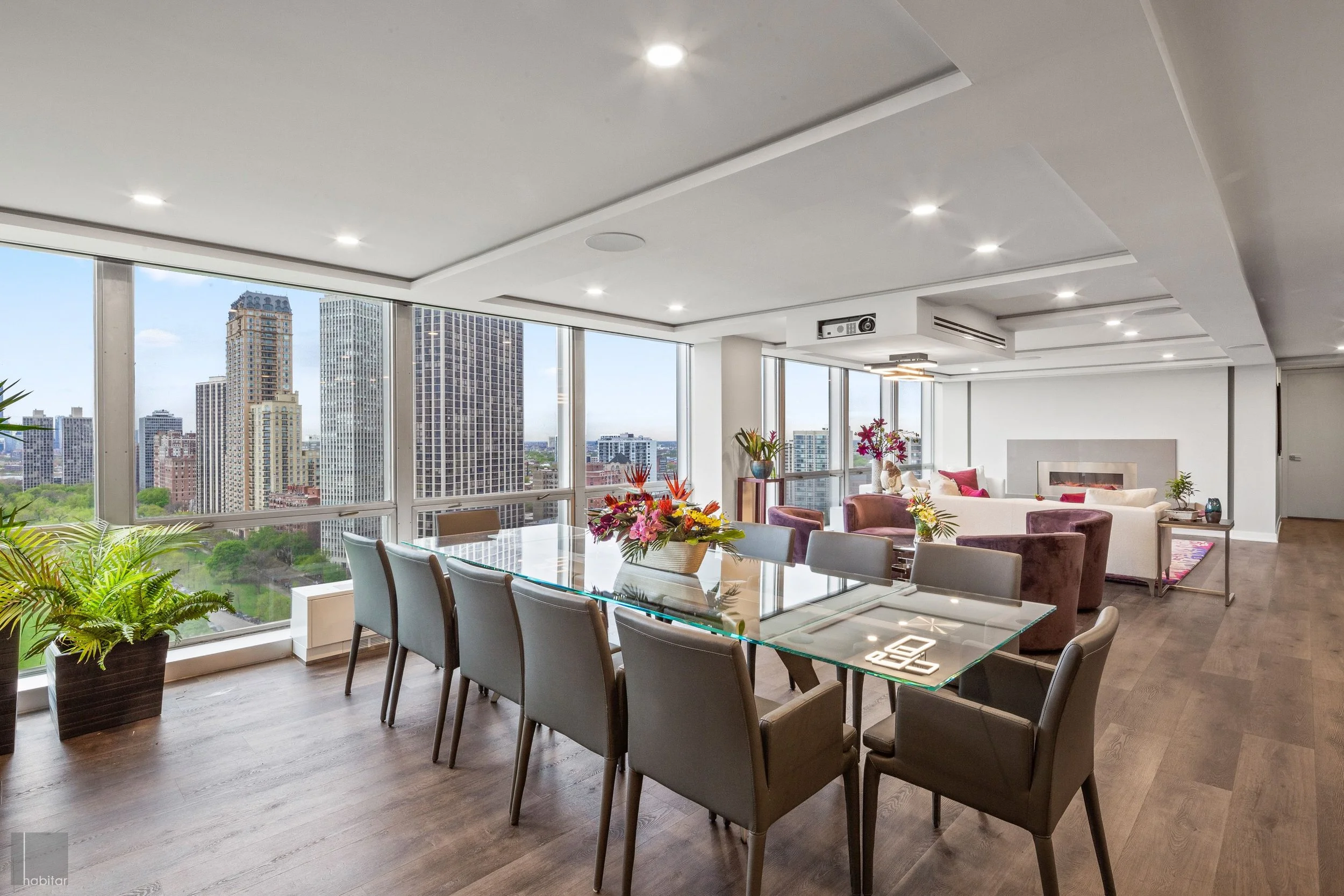 Modern apartment living and dining area with large windows showing city skyline, featuring a glass dining table with chairs and a spacious seating area with sofas, plants, and floral arrangements.