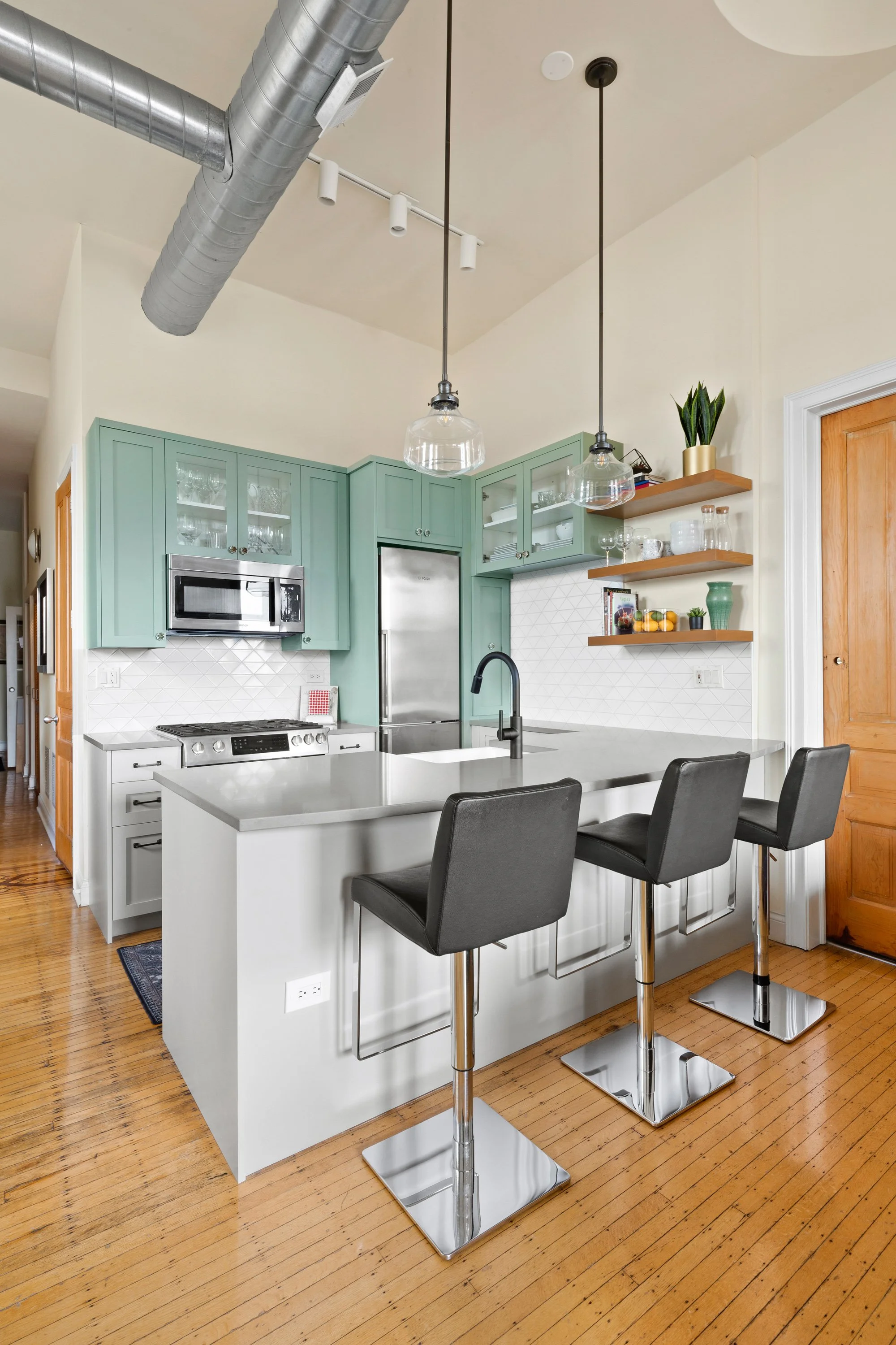 Modern kitchen with mint green cabinets, stainless steel appliances, white countertops, and black bar stools at a kitchen island, hardwood flooring, and open shelving with decorative items.