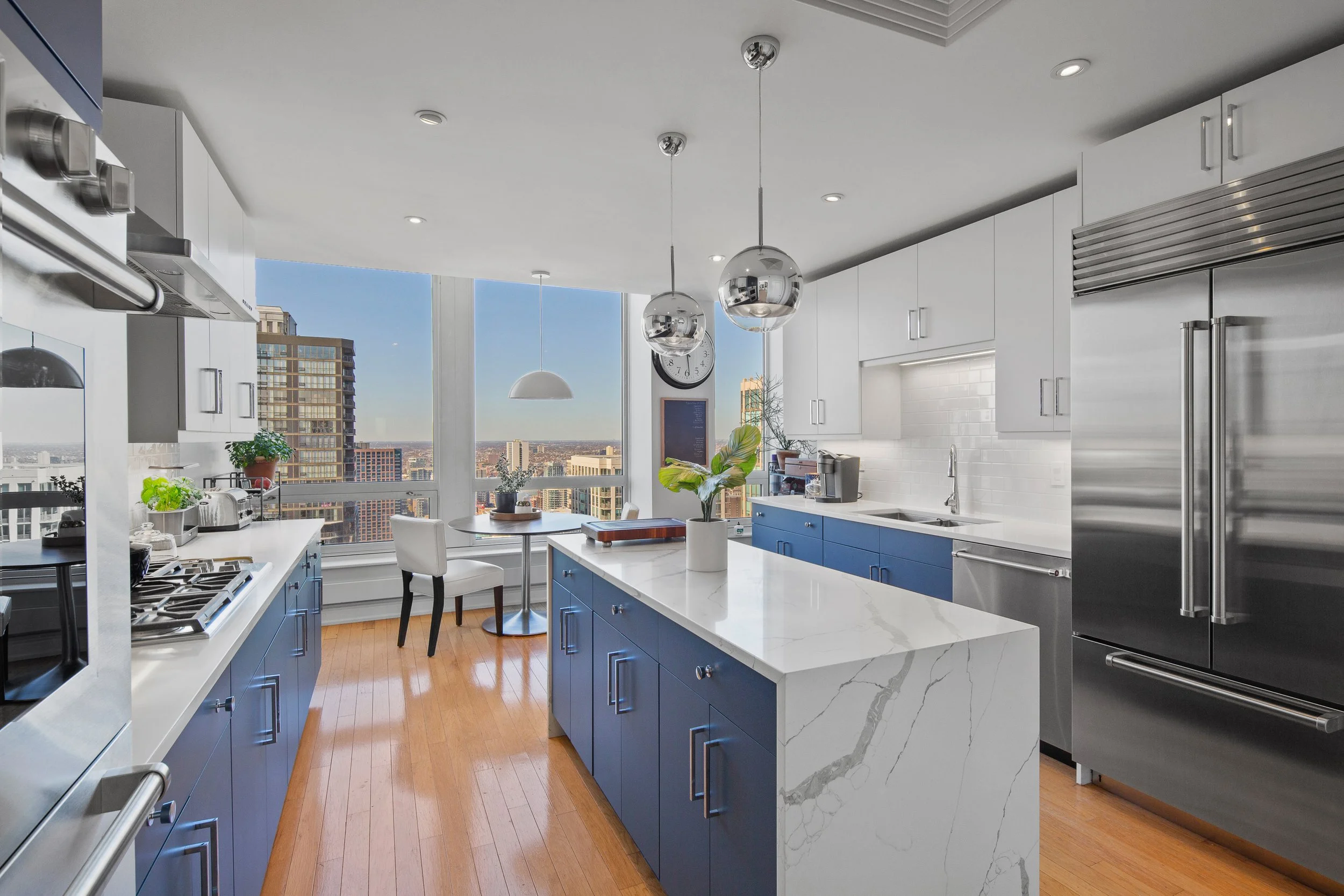 Modern kitchen with white cabinets with blue lower cabinets, a marble island, stainless steel appliances, wooden floors, a large window with a city view, hanging bulbs, a small dining table with chair by the window, plants, and a clock on the wall.