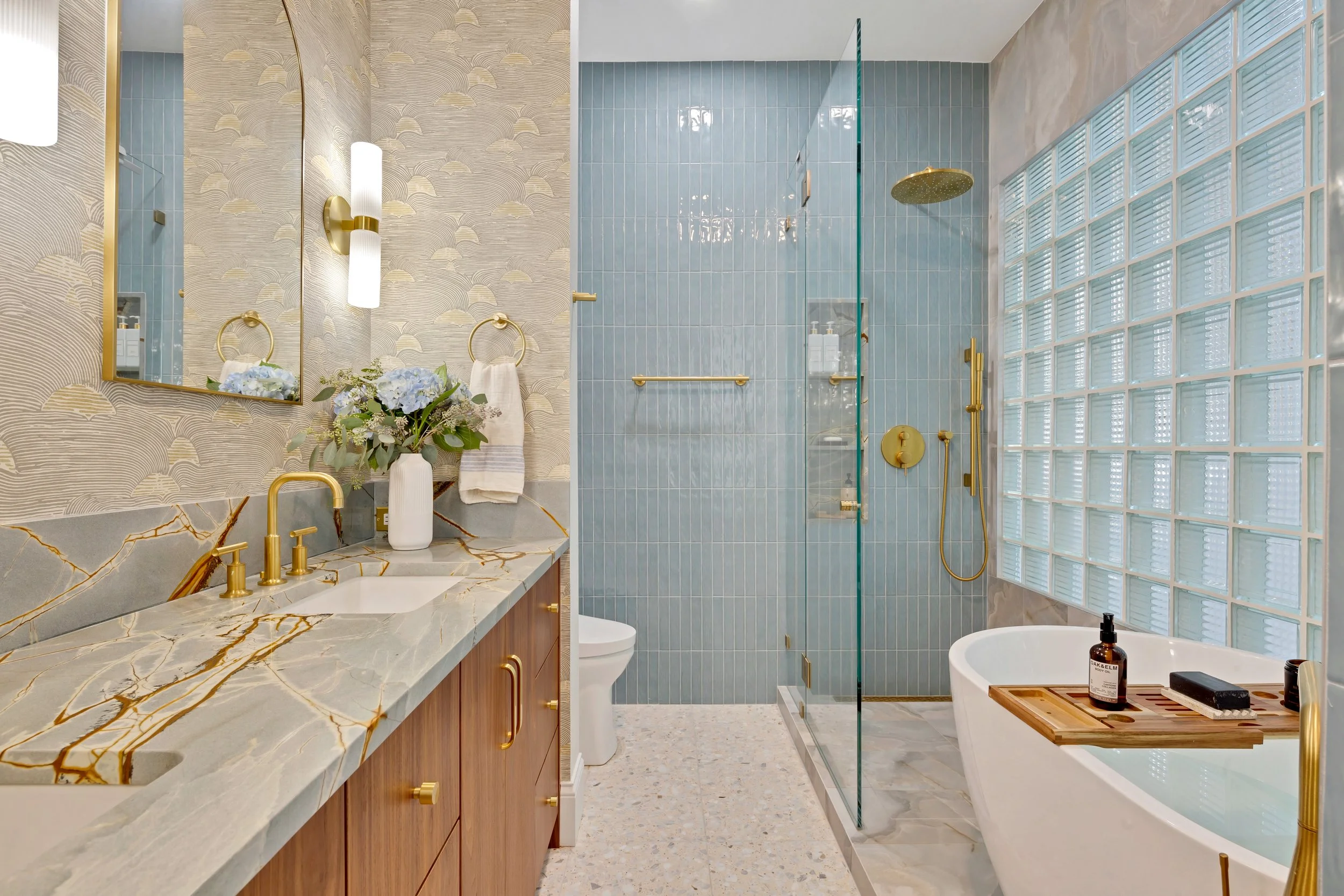 Luxurious bathroom with gold accents, blue tiled shower area, glass block wall, bathtub with bath tray, and marble countertop with a vase of flowers.