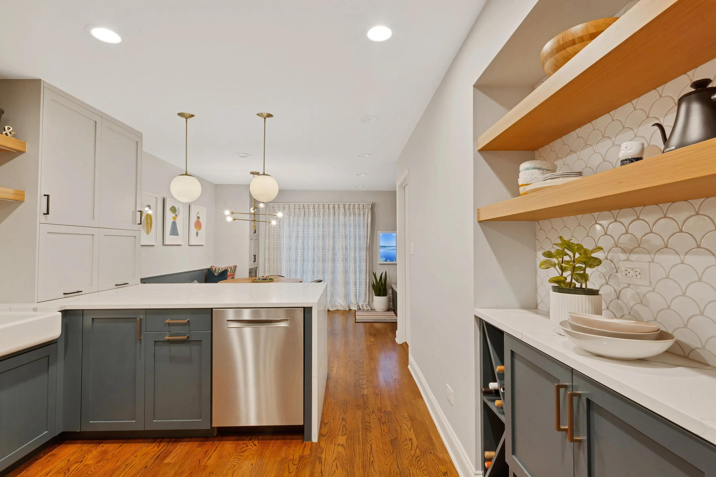Modern kitchen with gray cabinets, white countertops, and open wooden shelves displaying dishes and decor, hardwood flooring, overlooking a living area with curtains and wall art.