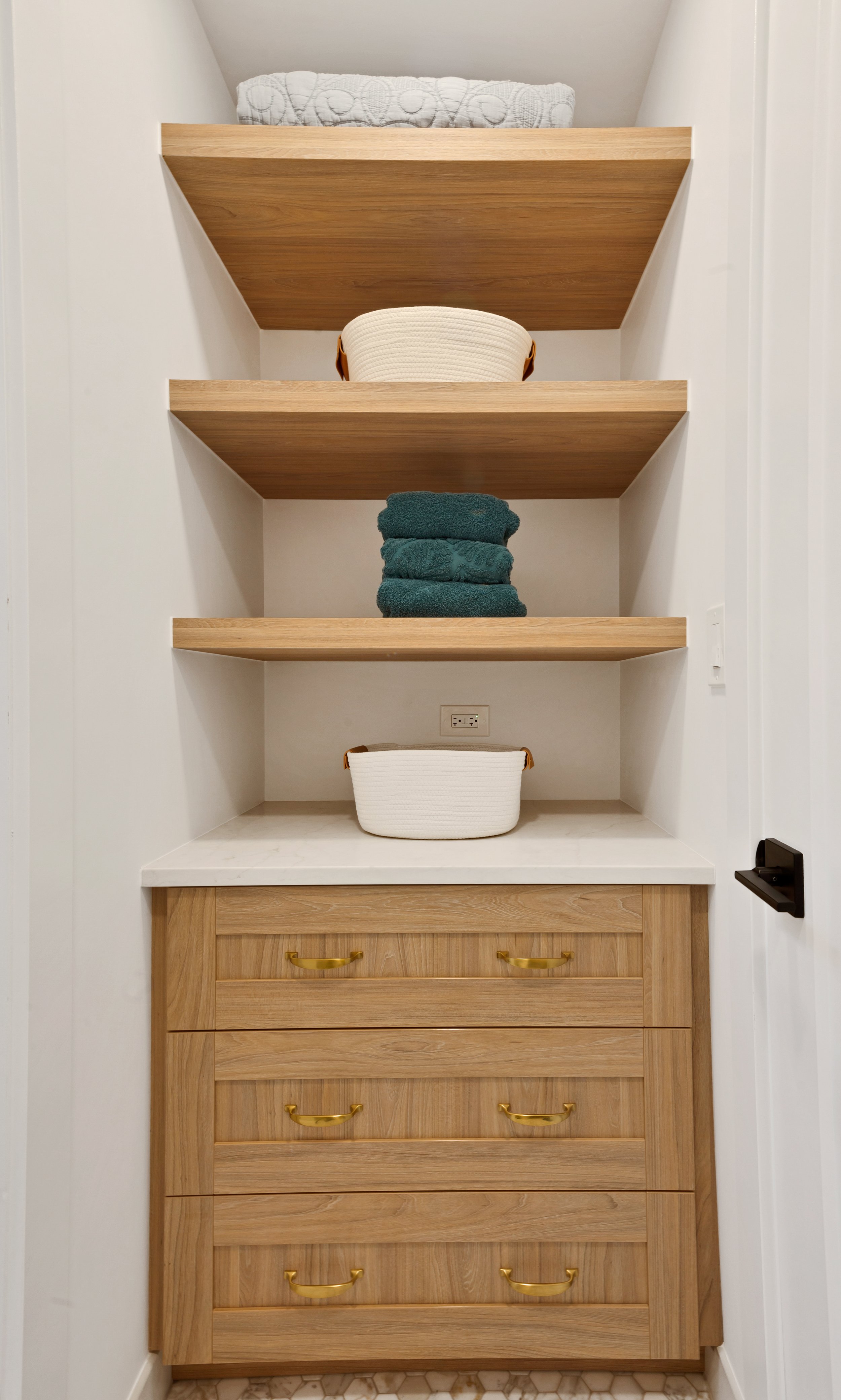 Built-in wooden shelving with towels, baskets, and linens, above a wooden cabinet with gold handles in a laundry or storage room.