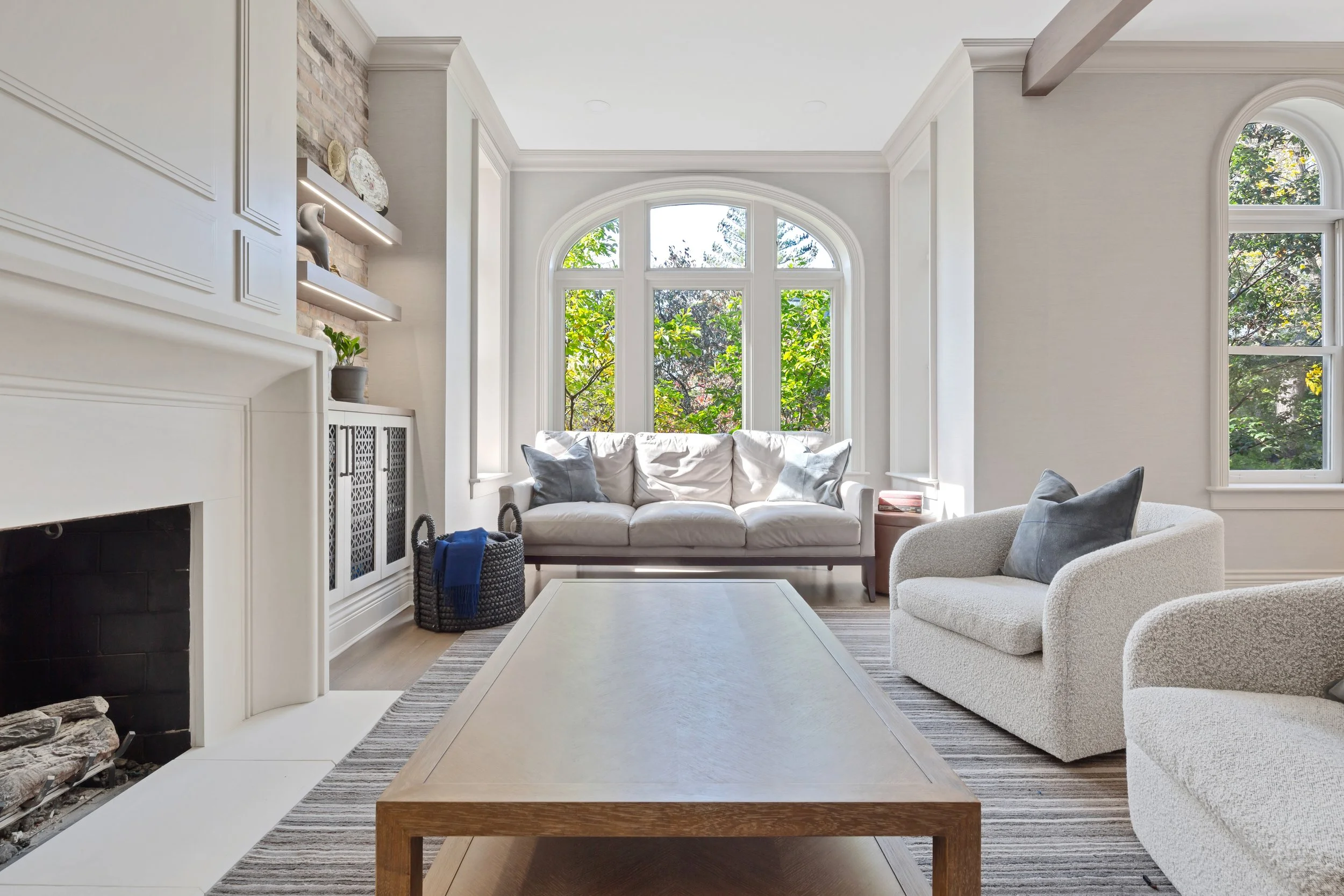 Bright living room with large arched window, white sofa with gray pillows, beige armchair, wooden coffee table, fireplace, and built-in shelves with decor, overlooking green trees outside.