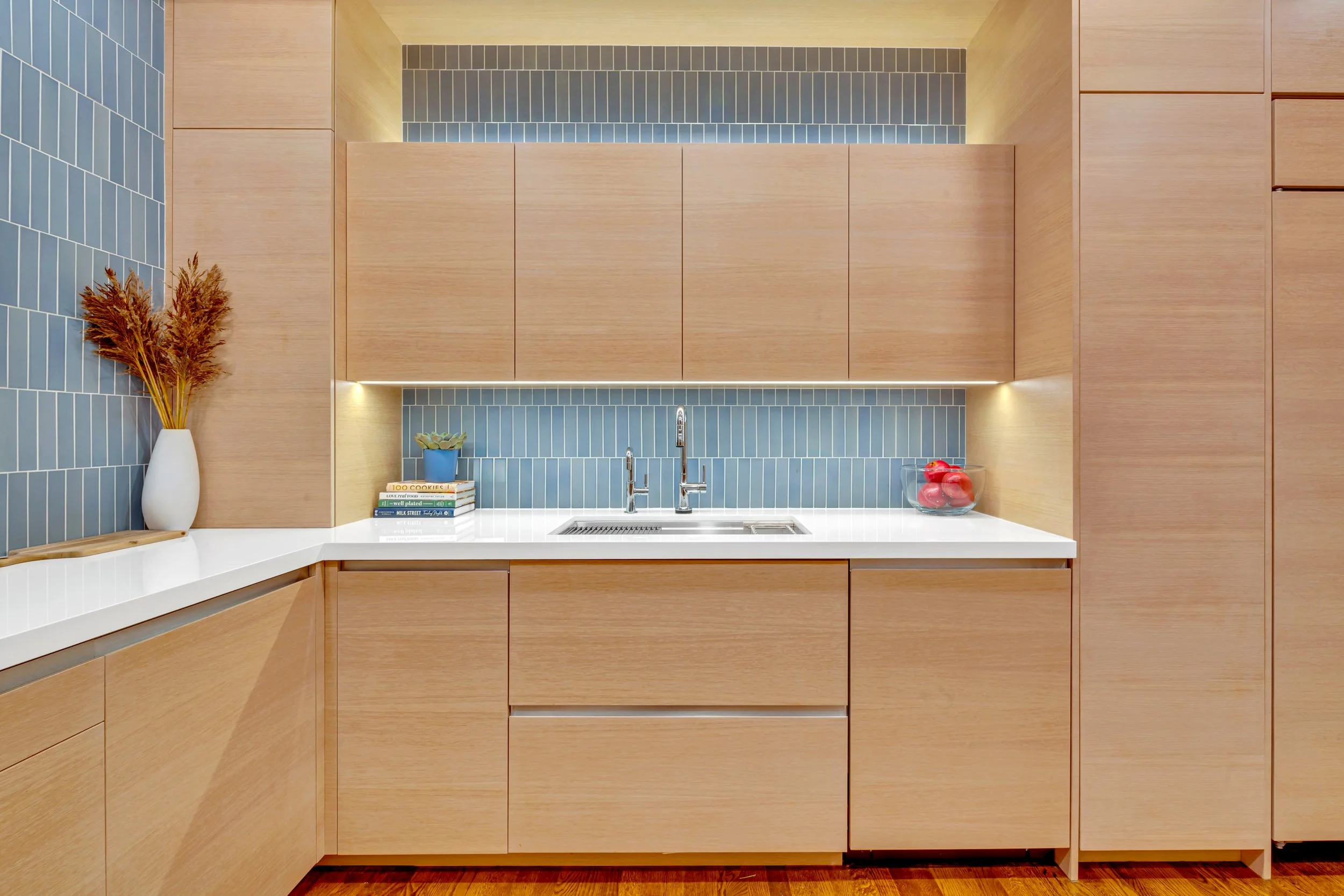 Modern kitchen with wooden cabinets, blue tiled backsplash, white countertop, and decorative items including a vase with dried plants, a bowl of red apples, and a small plant.