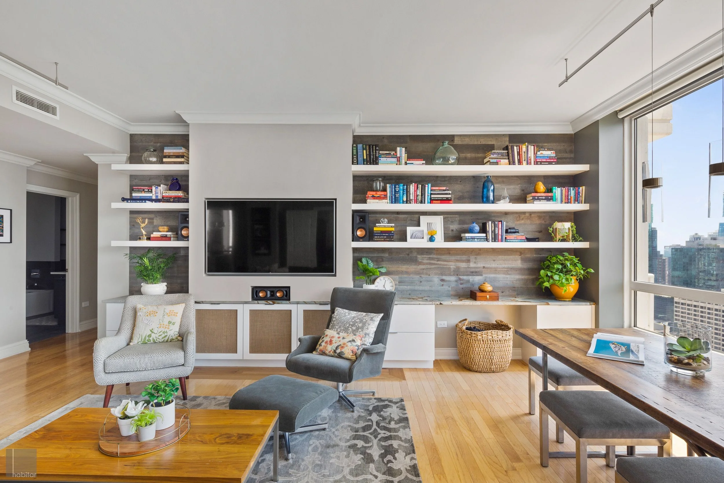 Living room with wooden floor, large window, bookshelves, TV, gray armchair with floral pillow, dining table, and houseplants.