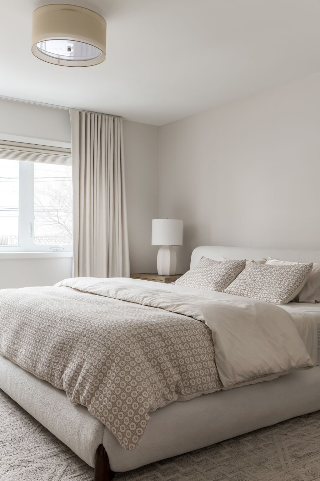 A neatly made bed with beige patterned bedding and matching pillows in a minimalist bedroom with a white headboard, bedside table with a white lamp, beige curtains, and a window with blinds.