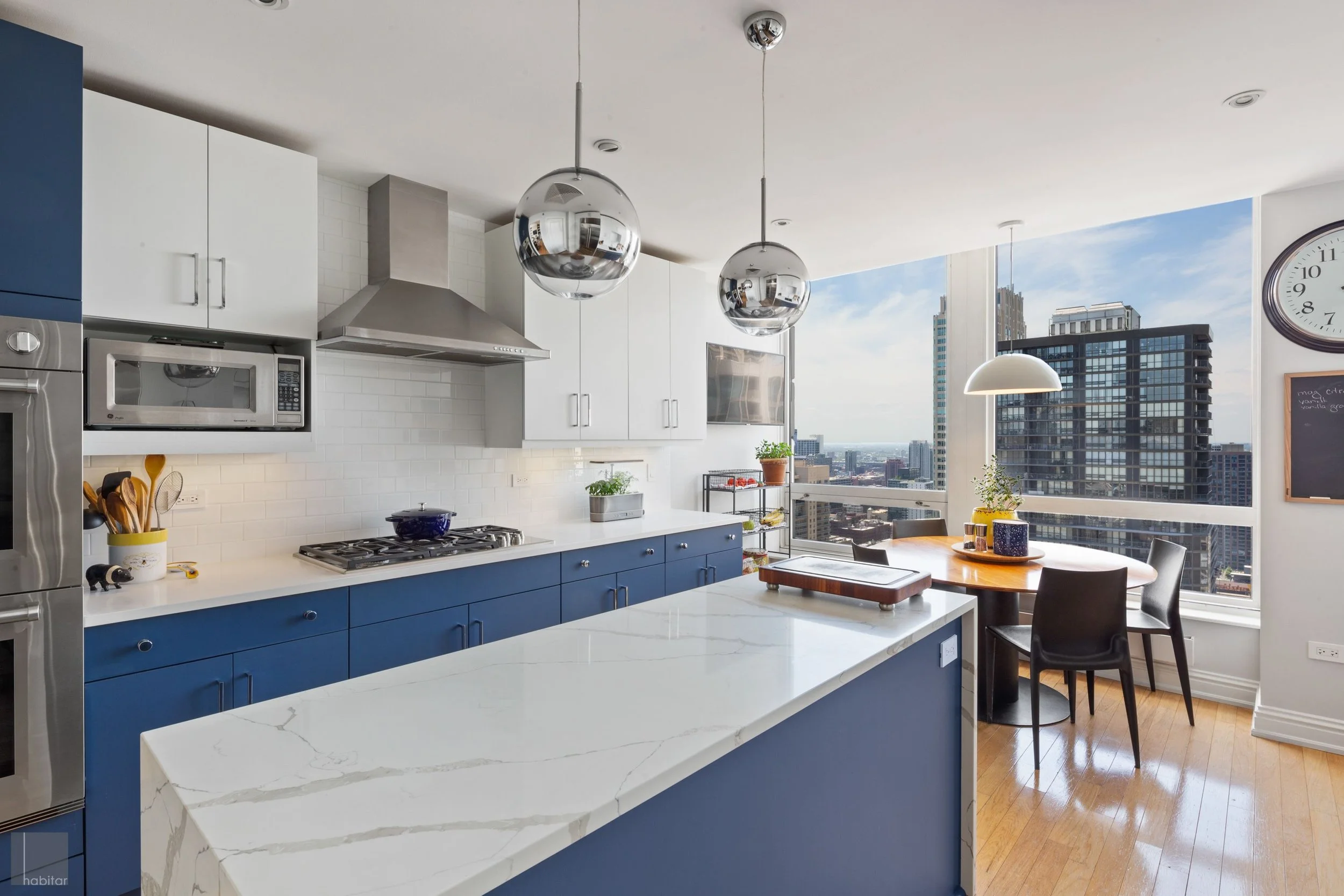 Modern kitchen with white and blue cabinets, marble island, stainless steel appliances, large windows with city view, round dining table with three chairs, and decorative plants.