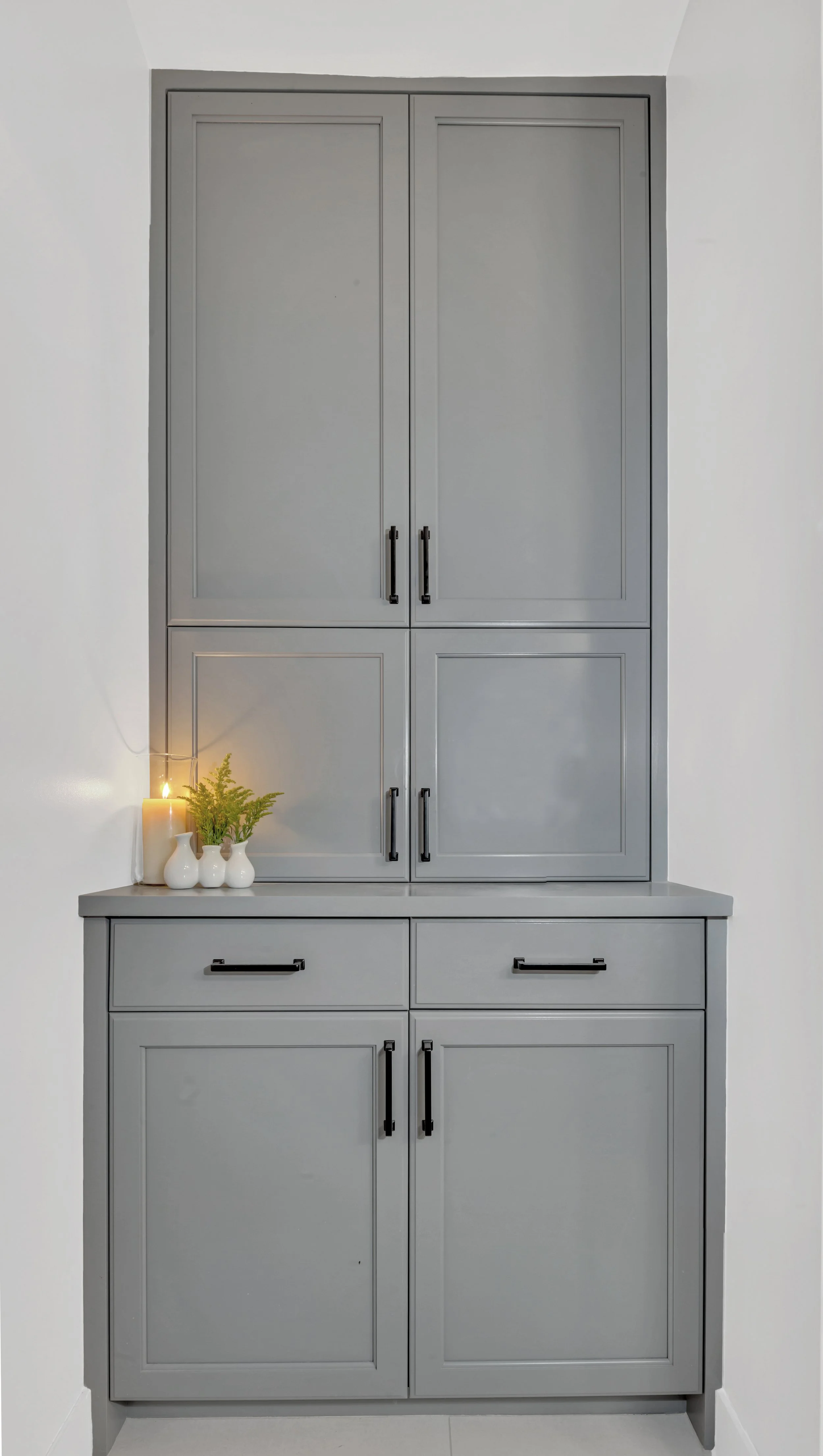 A built-in gray cabinet with black handles, organized in two sections with upper and lower doors, placed against a white wall. On the left side of the lower section, there are three white vases, a small potted plant, and a candle providing a soft glo