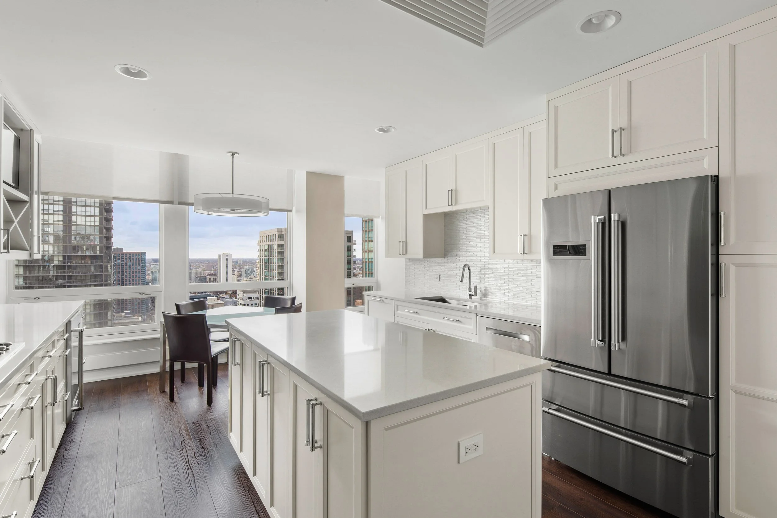 Modern kitchen with white cabinets, a large island, stainless steel appliances, and large windows overlooking a cityscape.