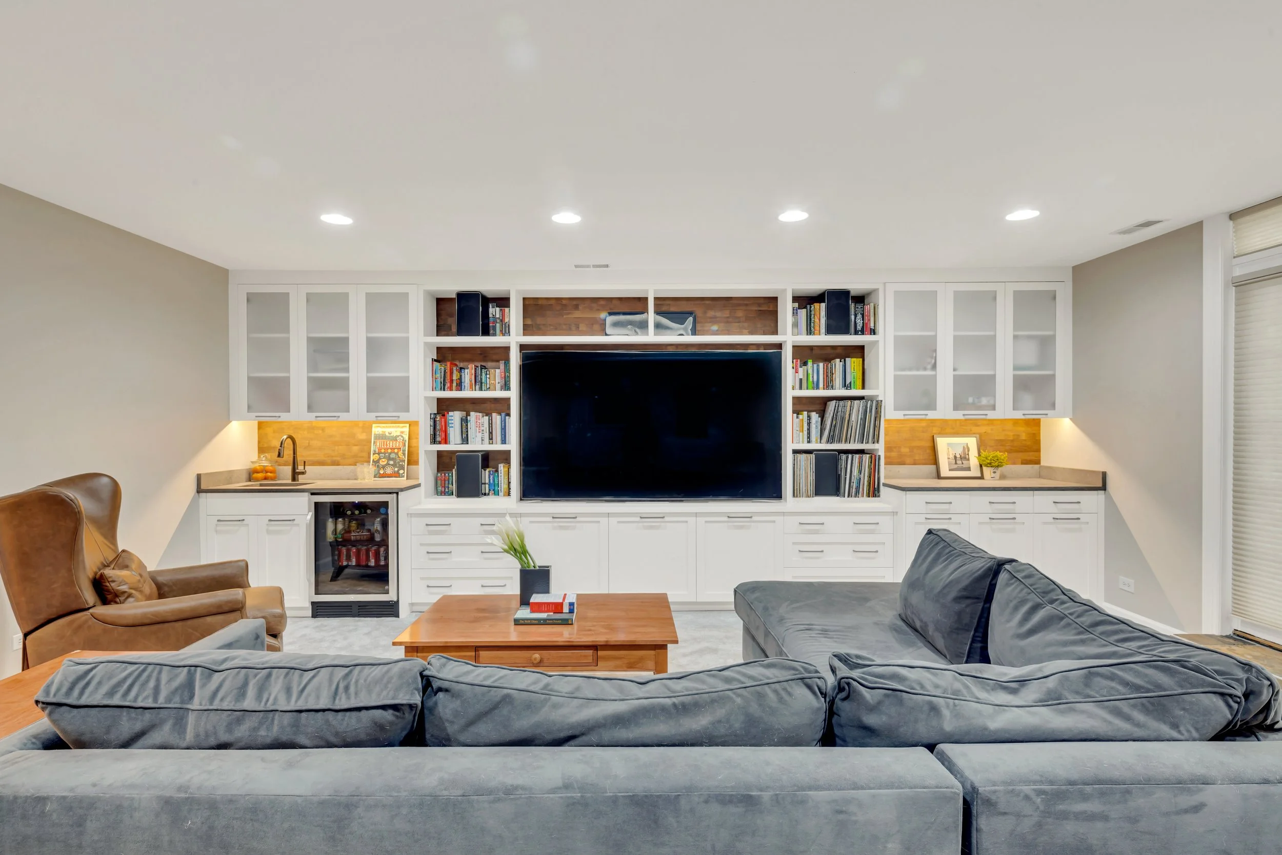 Modern living room with a large wall-mounted TV, white built-in shelves filled with books, a gray sofa, a wooden coffee table with books and a small plant, and a brown leather armchair.