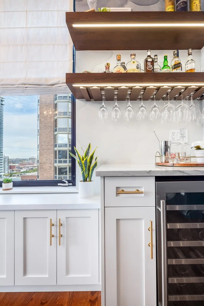 Modern kitchen with white cabinets, a potted plant on the countertop, a window with city view, and floating shelves holding bottles and hanging glassware.