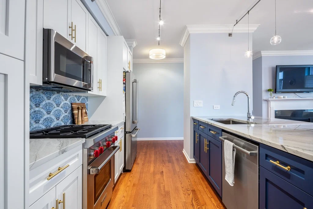 Modern kitchen with blue and white cabinets, hardwood floors, stainless steel appliances, and a marble countertop.