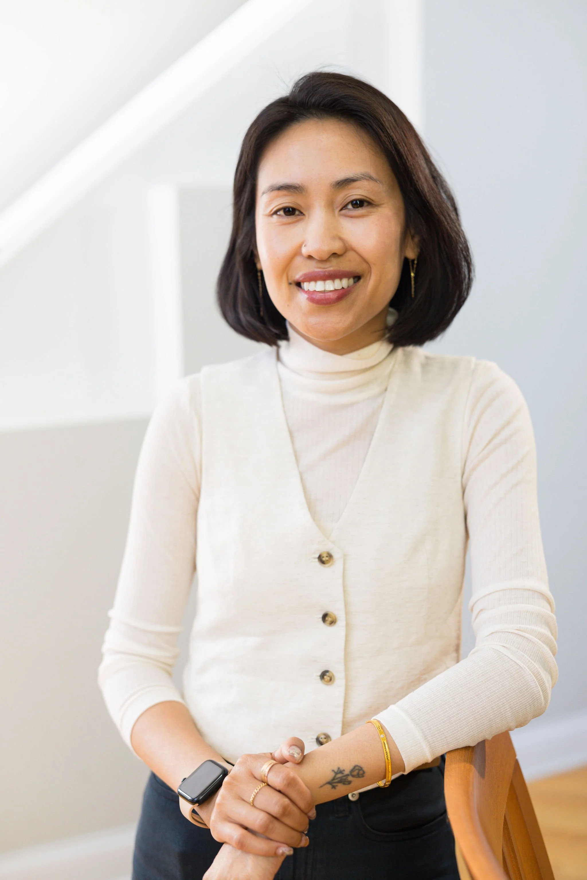 A woman with shoulder-length dark hair smiling at the camera, wearing a cream-colored turtleneck sweater, accessorized with earrings, bracelets, and a smartwatch.
