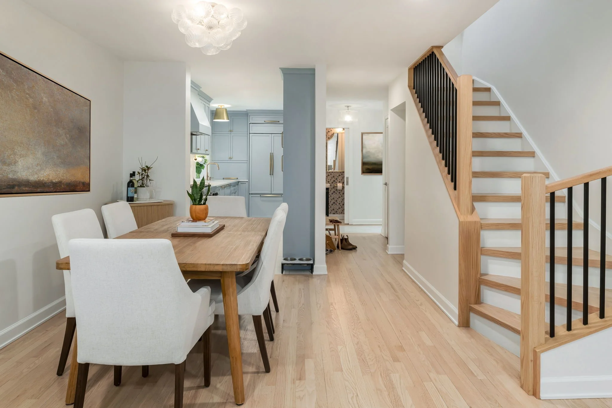 Interior view of a dining area with a wooden table and white upholstered chairs, adjacent to a staircase with wooden steps and black balusters, leading to the upper floor, with a kitchen in the background and a light fixture above.