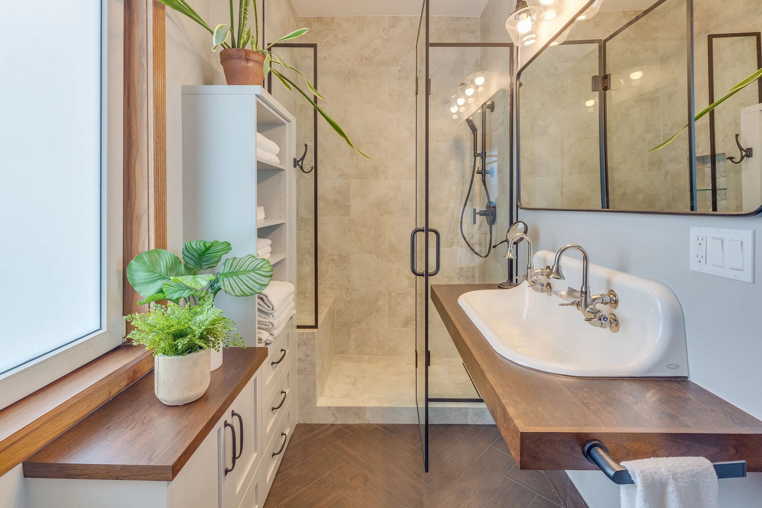 Modern bathroom with a walk-in shower, large mirror, wooden vanity with an elongated sink and multiple faucets, white shelf with towels, potted plants on a wooden countertop, and a window with natural light.