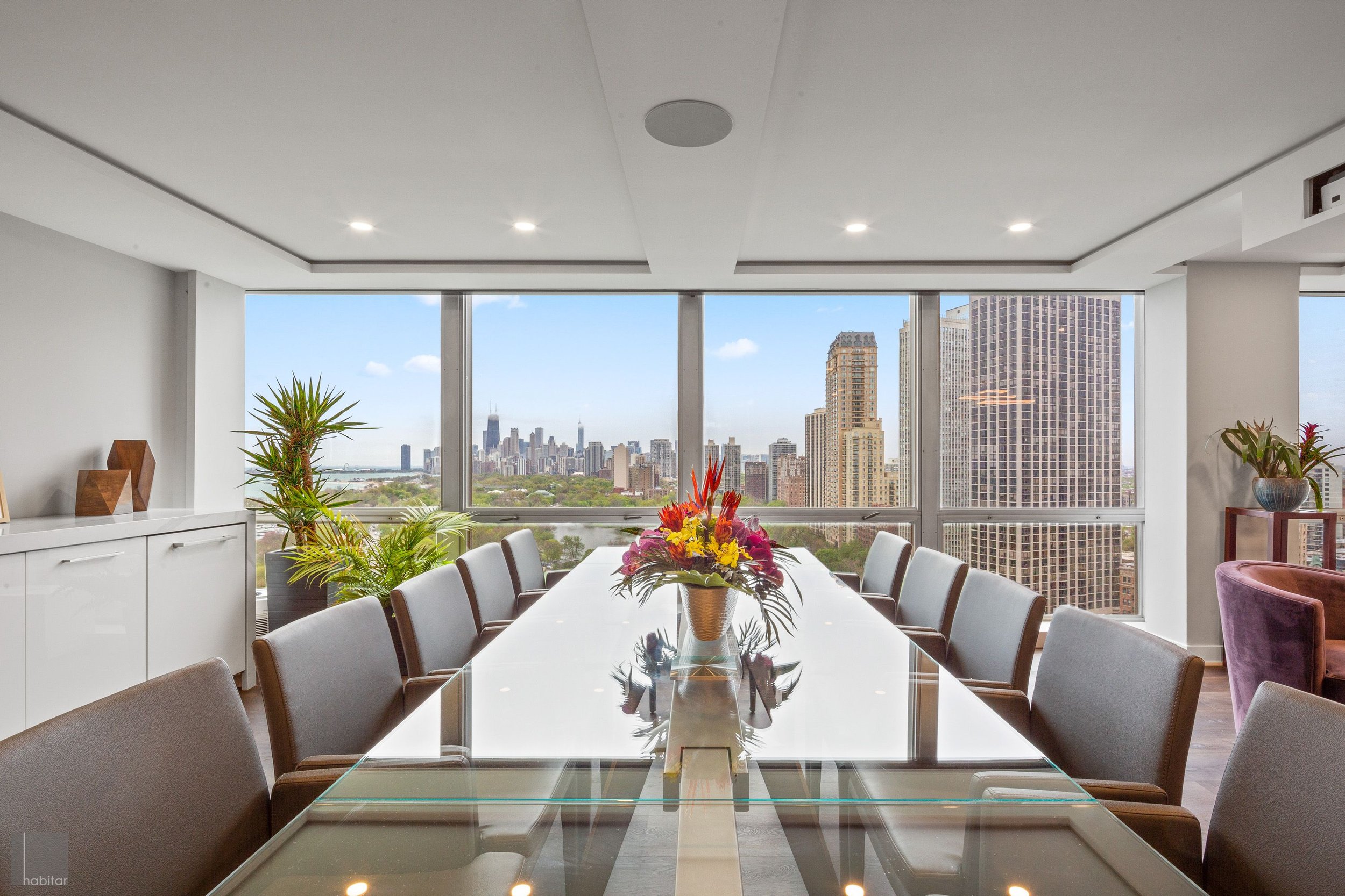 Modern conference room with a large glass table, surrounded by grey chairs, overlooking New York City skyline through large windows, decorated with potted plants and a colorful bouquet of flowers.