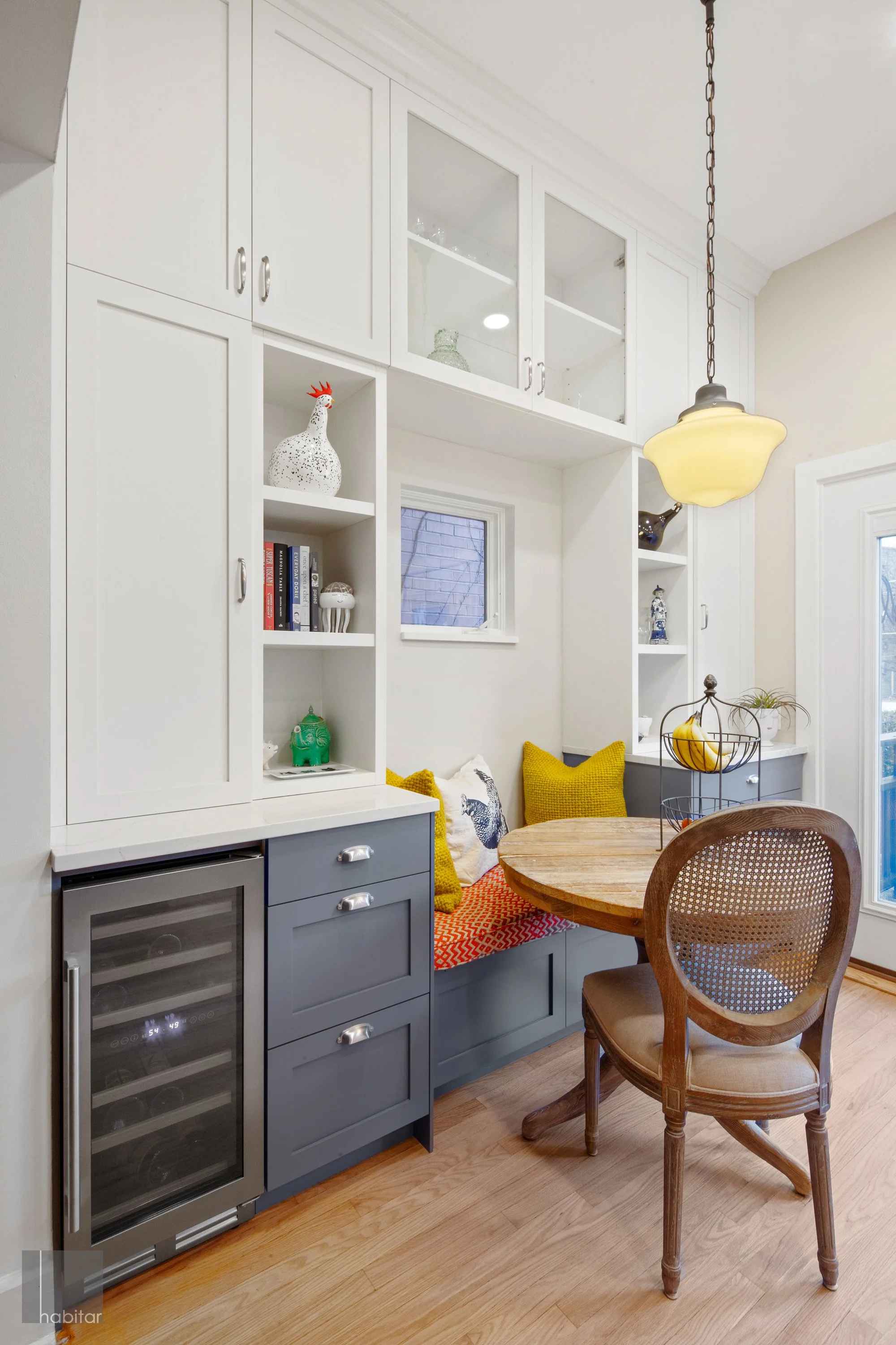 Kitchen nook with gray built-in benches, colorful cushions, a small round wooden table, a vintage wooden chair, and white cabinetry with glass and open shelves.