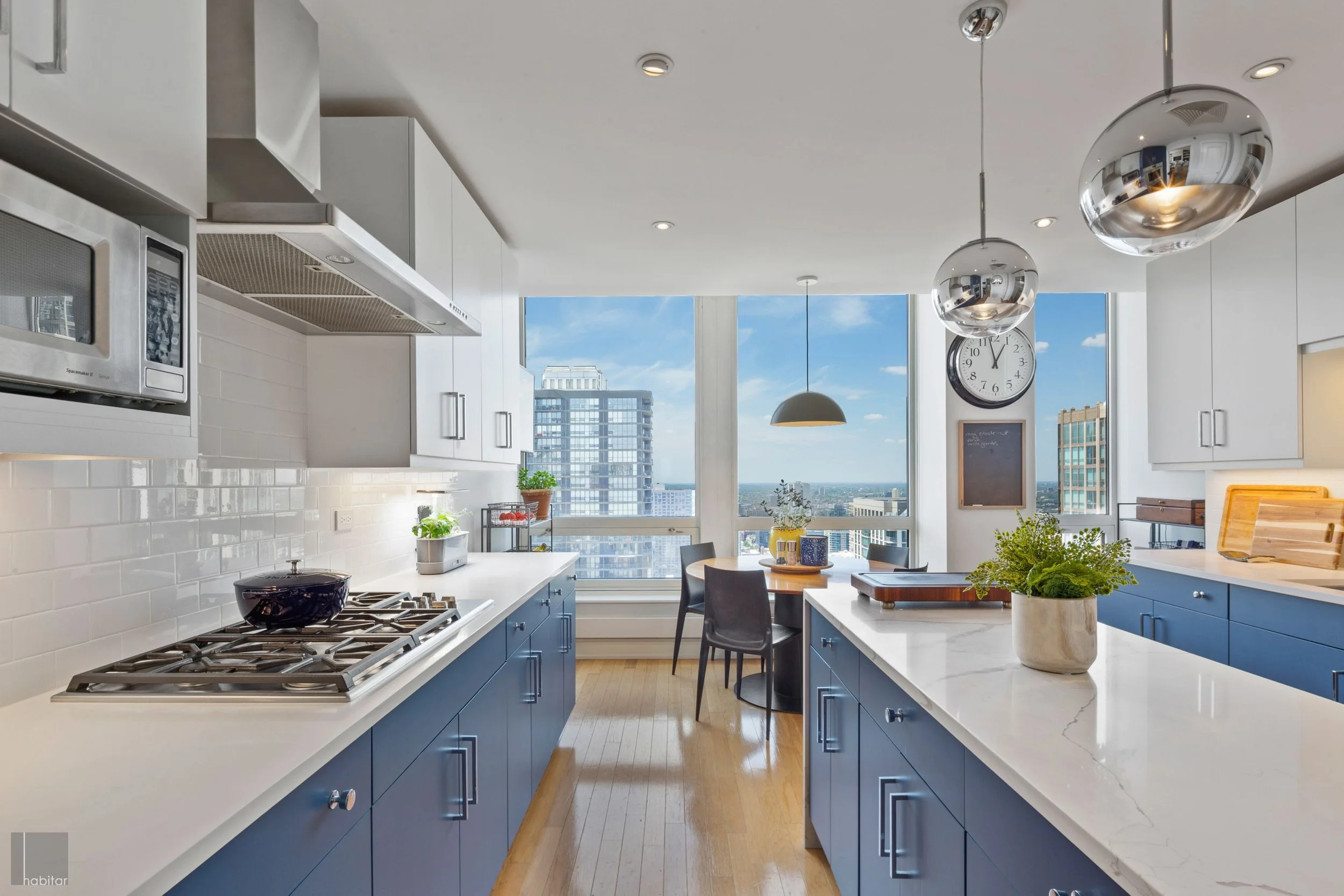 Modern kitchen with blue cabinets, white countertops, stainless steel appliances, and large windows showing a cityscape view.