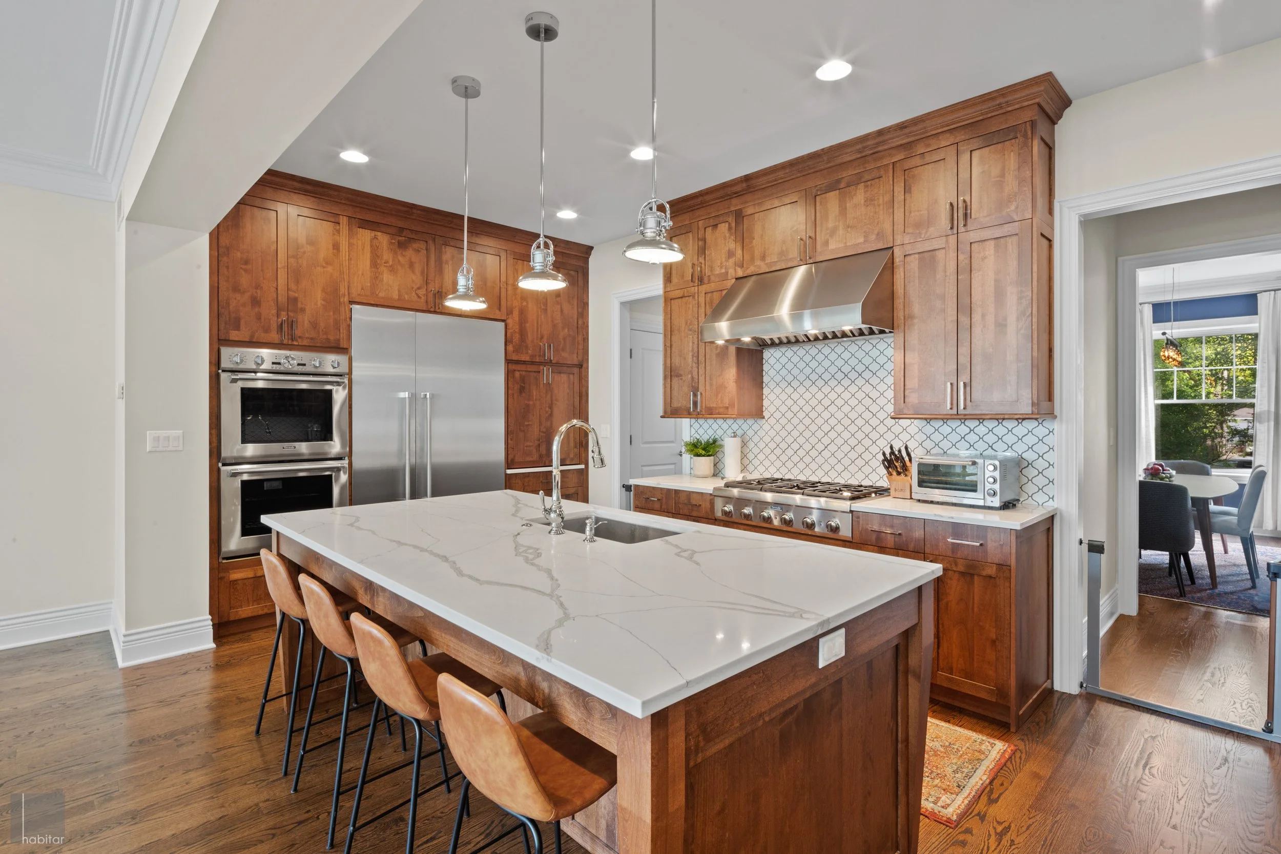 Modern kitchen with wooden cabinets, a white marble island with a sink and seating, stainless steel appliances, and a backsplash with a geometric tile pattern.
