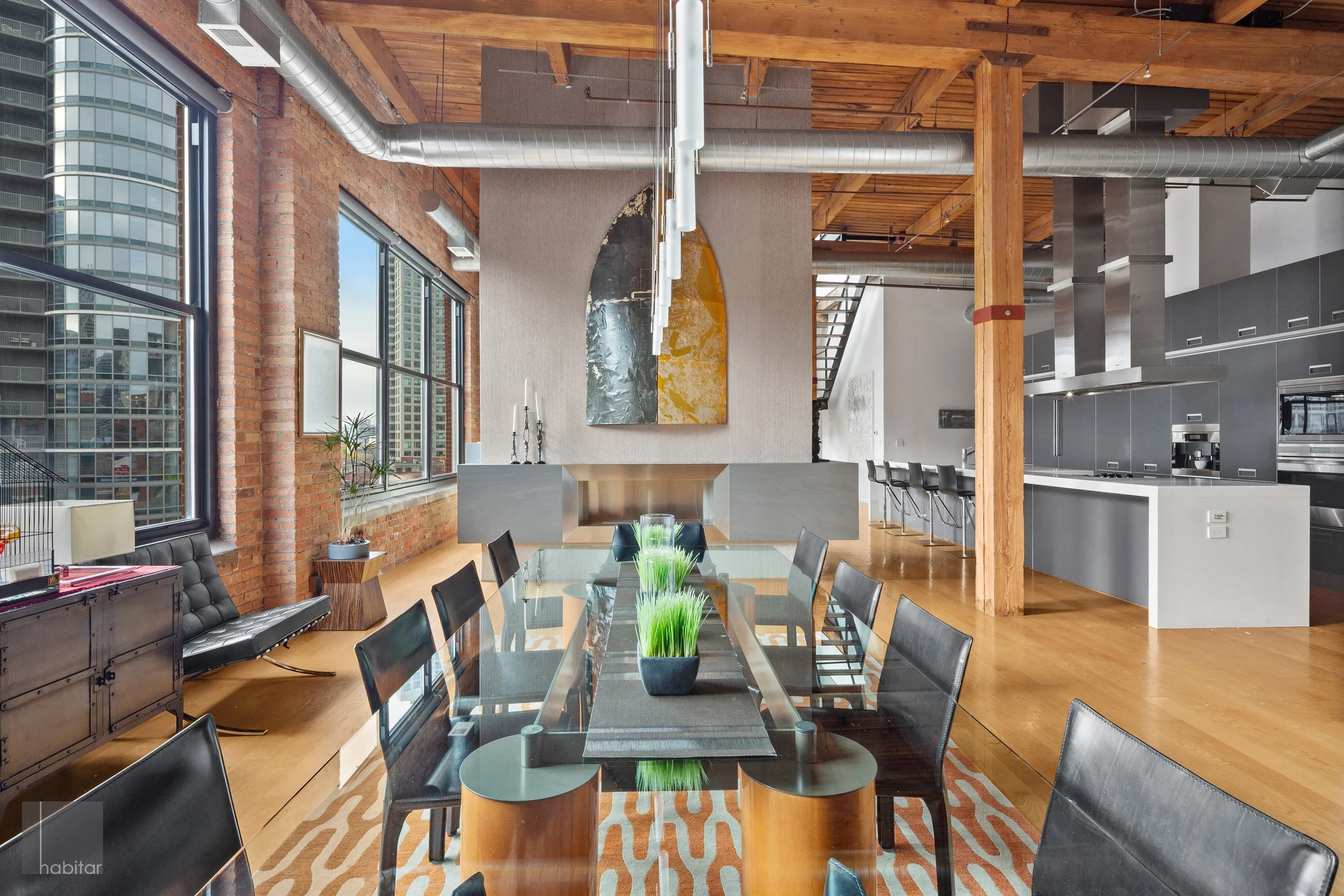 Modern loft-style dining area with large windows, wooden beams, and exposed brick walls. Features a glass dining table with black chairs, a decorative wall mirror, and an open-concept kitchen with stainless steel appliances.
