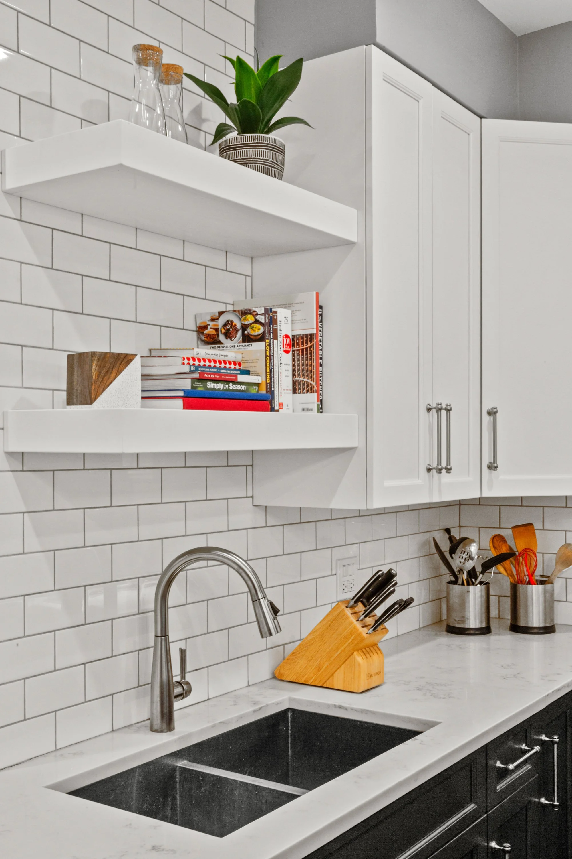 White kitchen with subway tile backsplash, open shelves with glass bottles, potted plant, cookbooks, and a black and white cabinet, with a stainless steel faucet, wooden knife holder, and utensils in containers on the marble countertop.