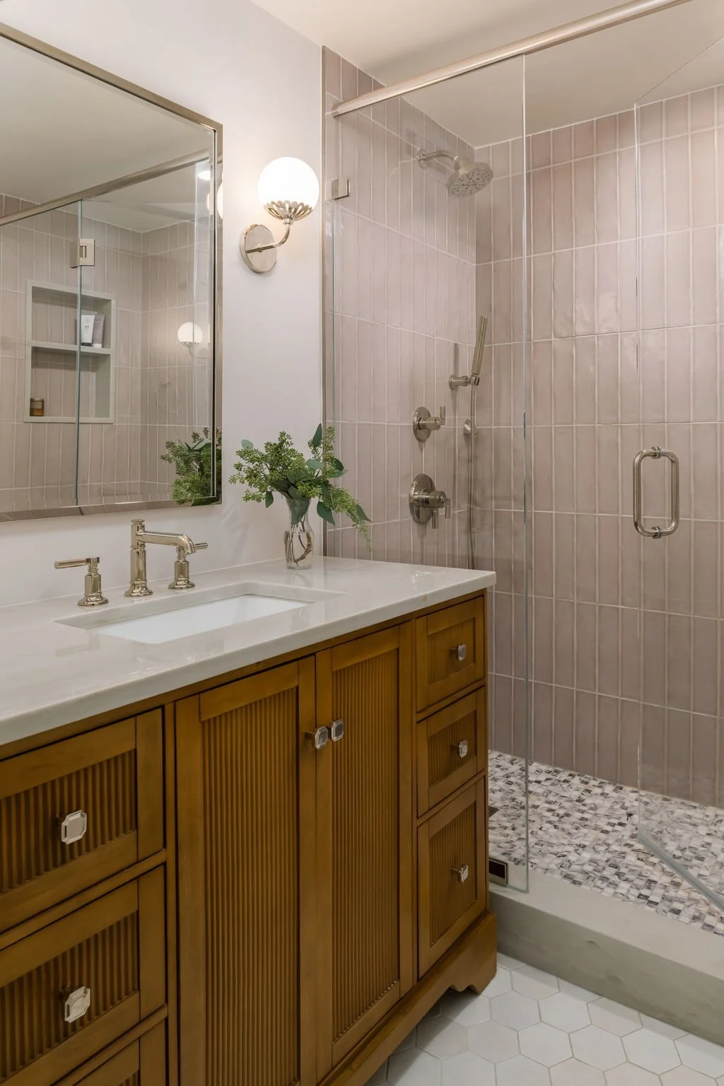 A bathroom with a wooden vanity, white countertop, and a rectangular mirror. There is a wall-mounted sconce light and a window reflecting in the mirror. The shower area has pink tiles, a glass door, and fixtures, including a showerhead and a handheld