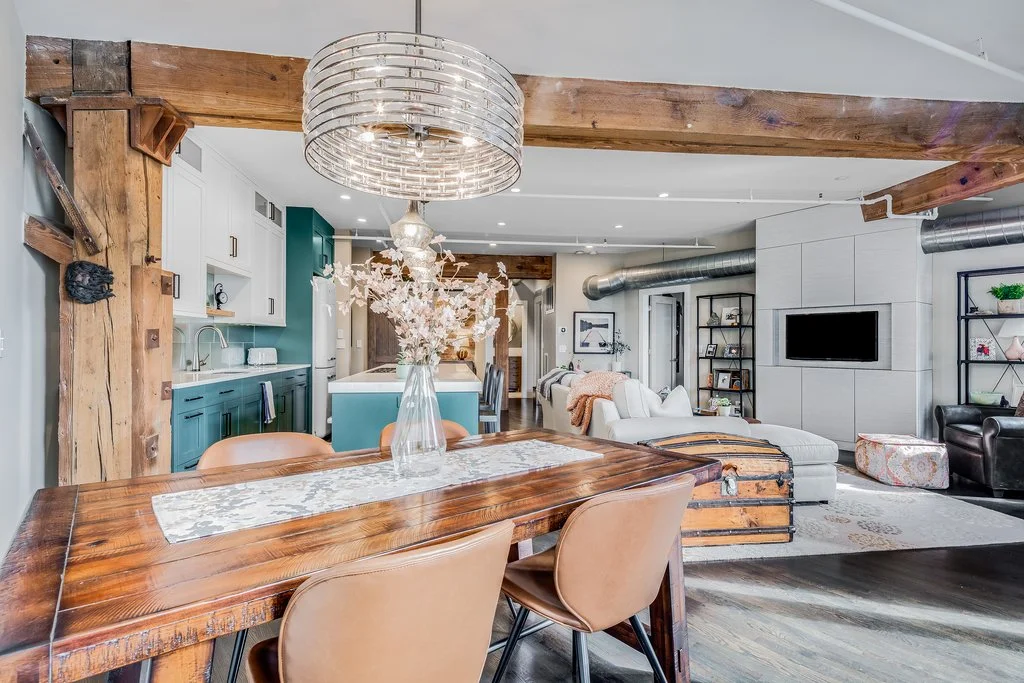 Open-concept living and dining area with a wooden dining table, tan chairs, a white and teal kitchen, exposed wooden beams, a chandelier, a white sectional sofa with a trunk coffee table, a wall-mounted TV, and black metal shelving units with decorat