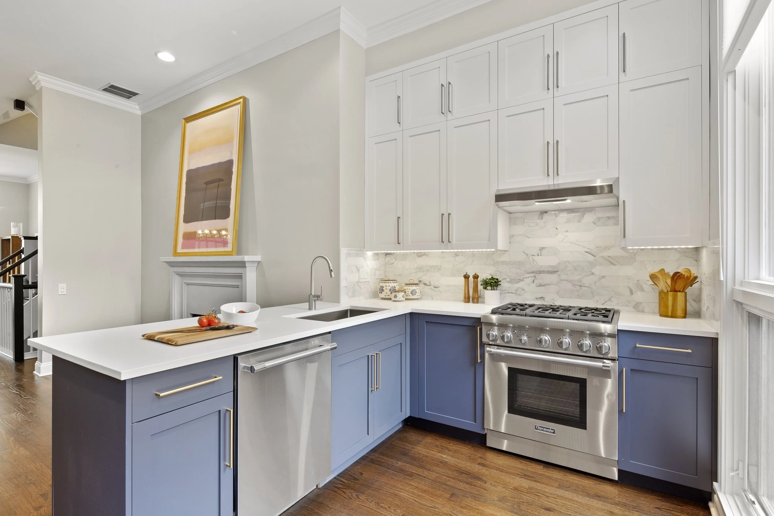 Modern kitchen with white upper cabinets, blue lower cabinets, stainless steel appliances, and a marble backsplash. A kitchen island with a white countertop, a sink, and a cutting board with tomatoes. Wooden flooring and a window on the right.