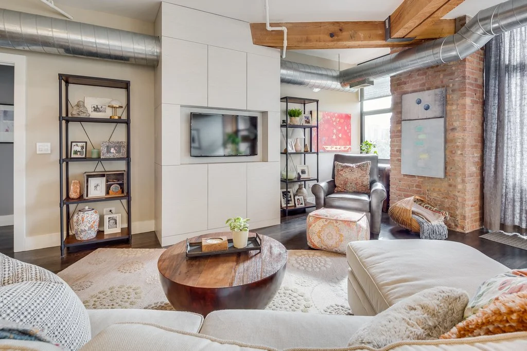 Cozy living room with beige sectional sofa, round wooden coffee table, black leather armchair, exposed brick wall, metal shelves with decorations, and artwork by window.