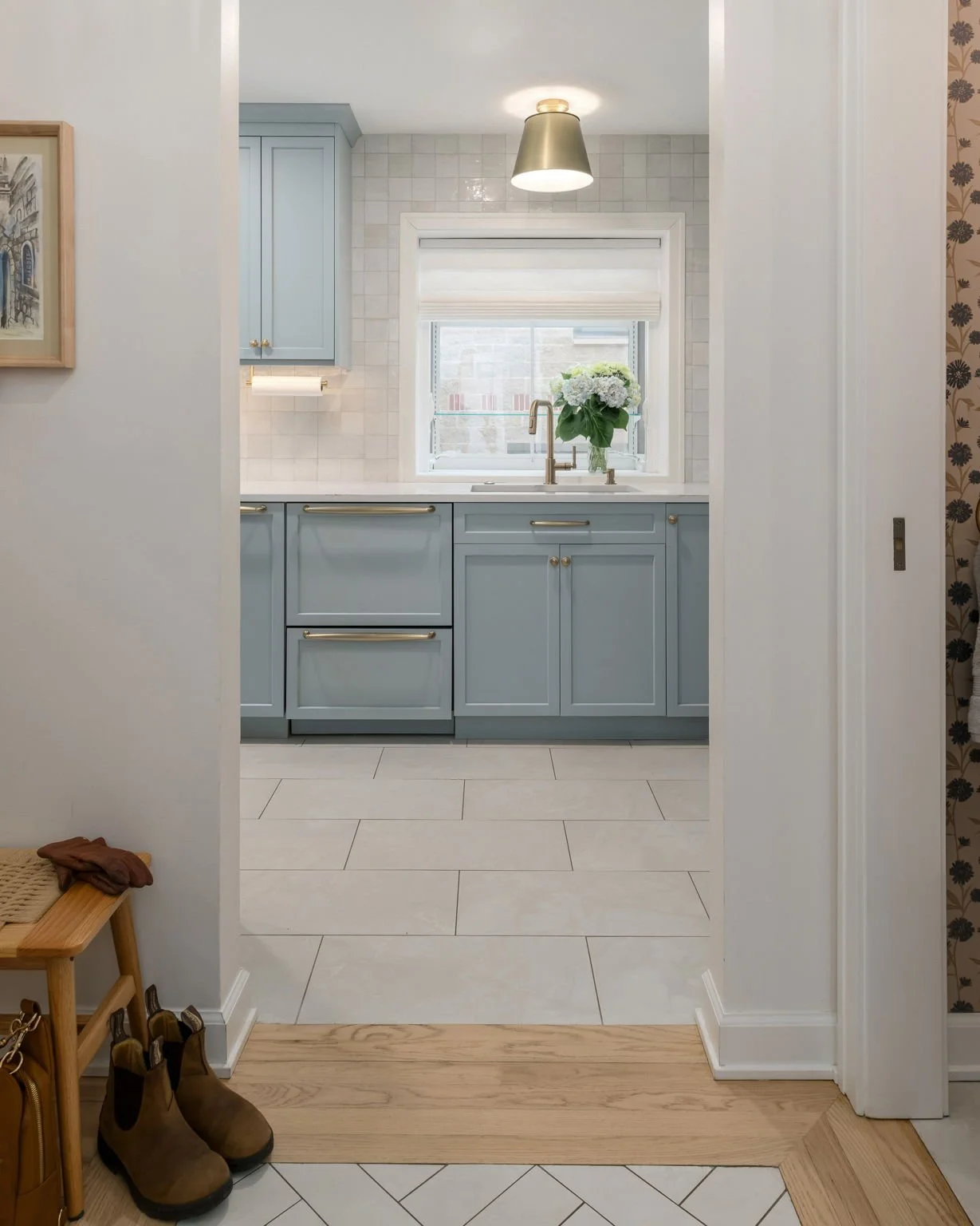 View of a kitchen with light blue cabinets, white tiled backsplash, window with a flower arrangement, and a ceiling light fixture.
