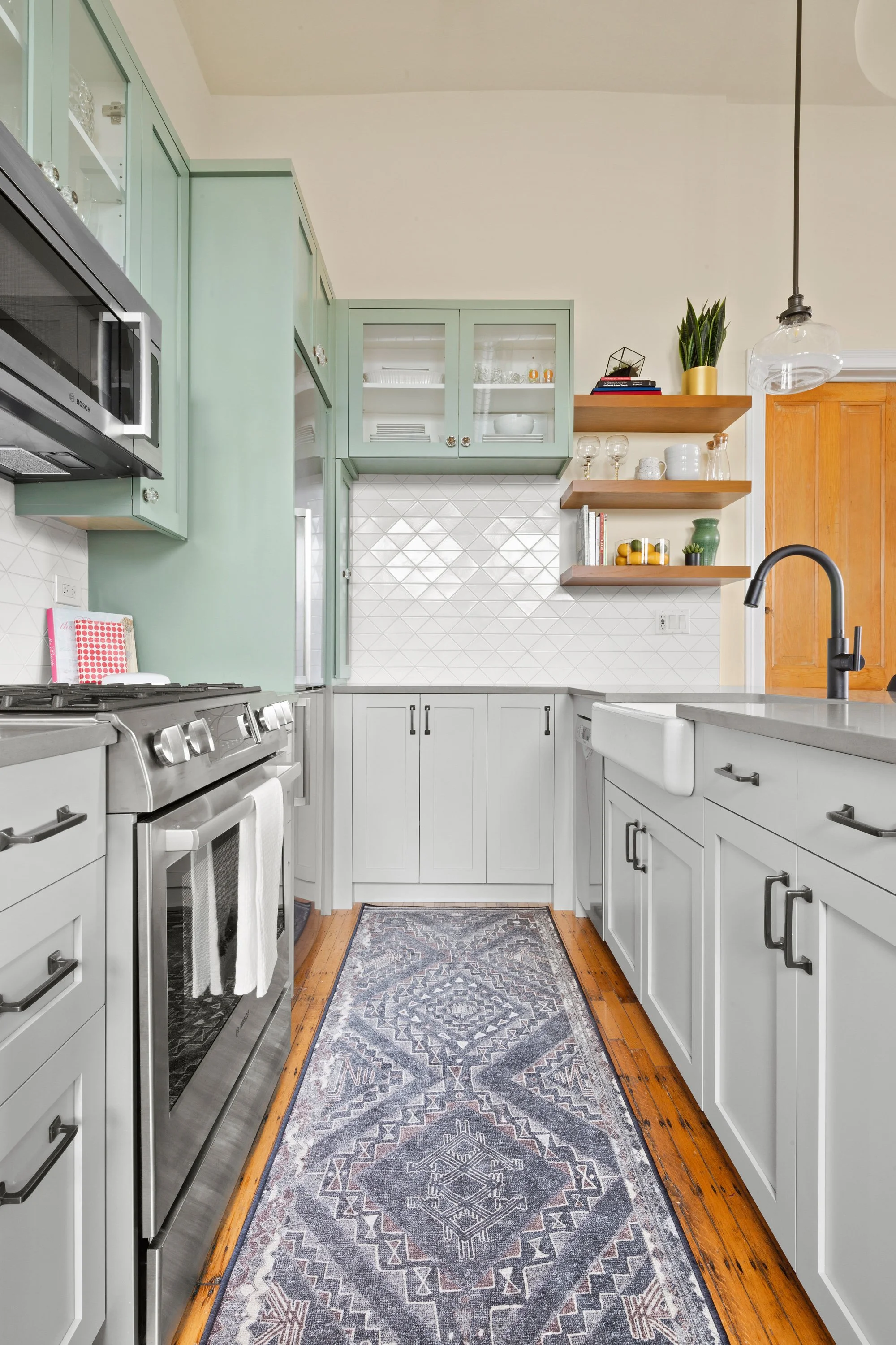 A modern kitchen with white cabinets, a stainless steel oven, green upper cabinets, open wooden shelves with decor, a black faucet, a patterned runner rug, and wooden flooring.