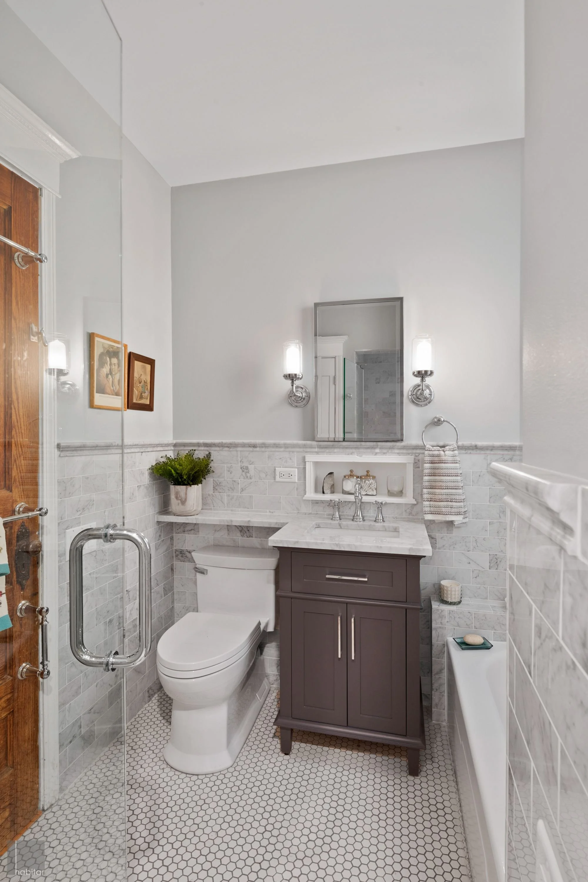 Bathroom with white tiles, dark vanity with marble countertop, mirror, wall-mounted lights, toilet, small plant, towel, and shower door.
