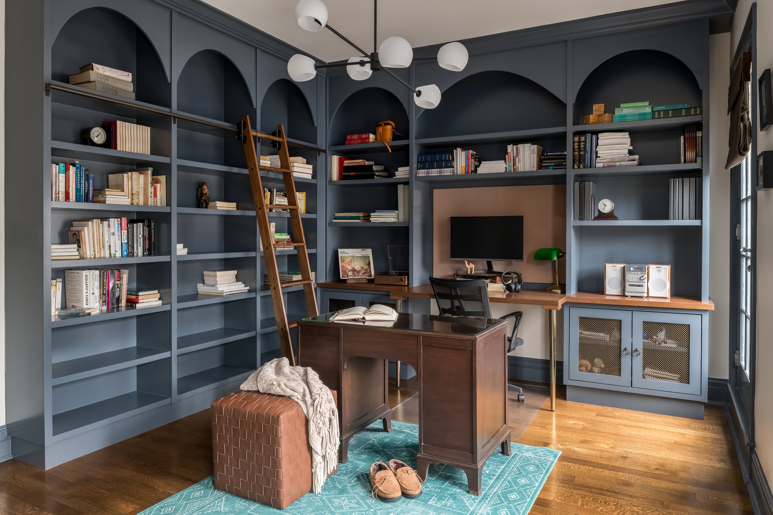 Room with blue built-in bookshelves, a wooden ladder, a desk with a computer monitor, a black office chair, a brown leather ottoman, a small turquoise rug, a green desk lamp, and various books and decorations on the shelves.