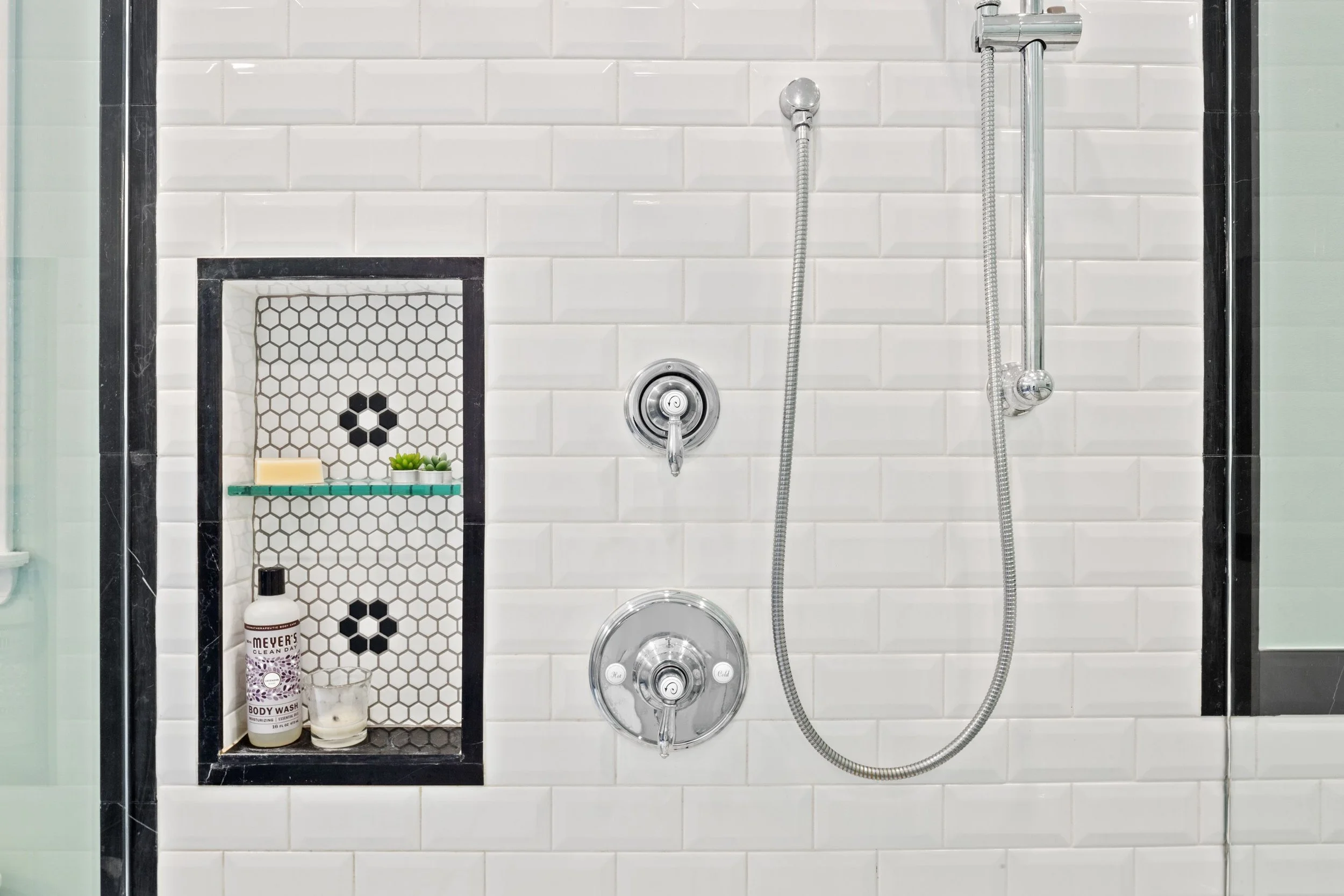 A modern shower area featuring white subway tiles, a handheld showerhead with a hose, and a circular control valve. There is a built-in niche with black hexagon and white tiles, containing soap, a small plant, a bottle of body wash, and a candle.