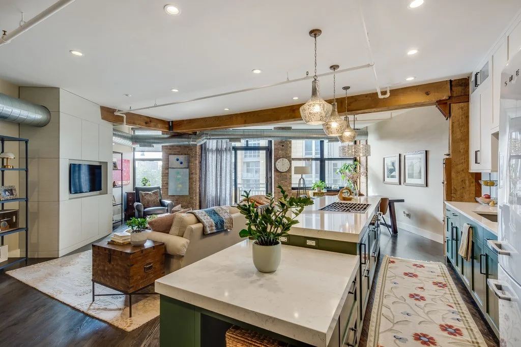 Open concept living room and kitchen with wooden beams, pendant lighting, a white couch, armchair, TV, shelving, large windows, potted plants, and a kitchen island with a plant on top.
