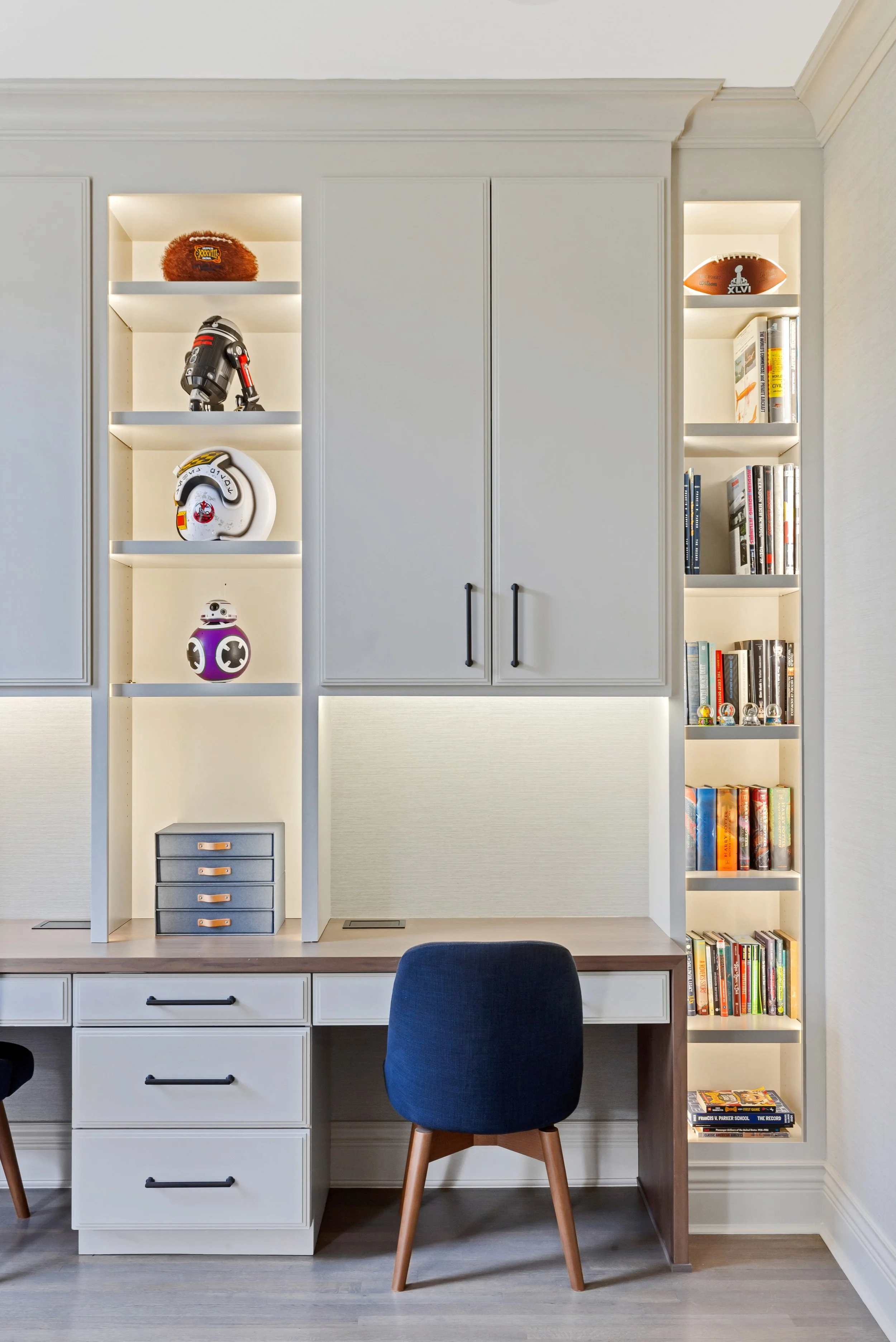 Home office desk with black chair and built-in white shelves containing books and collectibles, including Star Wars droids, helmets, and sports memorabilia.