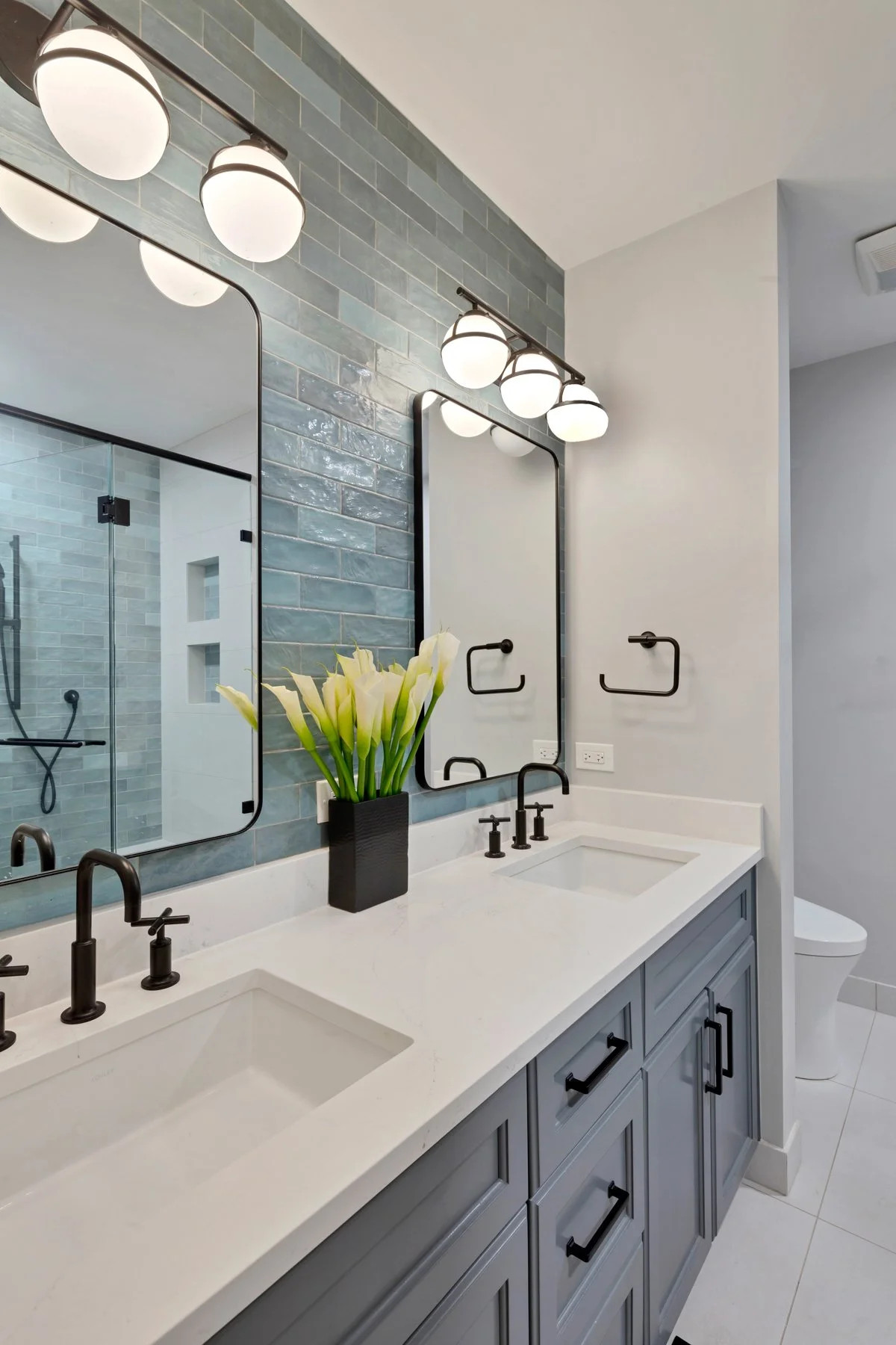 Bathroom with a double vanity, black fixtures, large mirrors, a blue tiled wall, a vase of white flowers, and a shower area with glass doors.