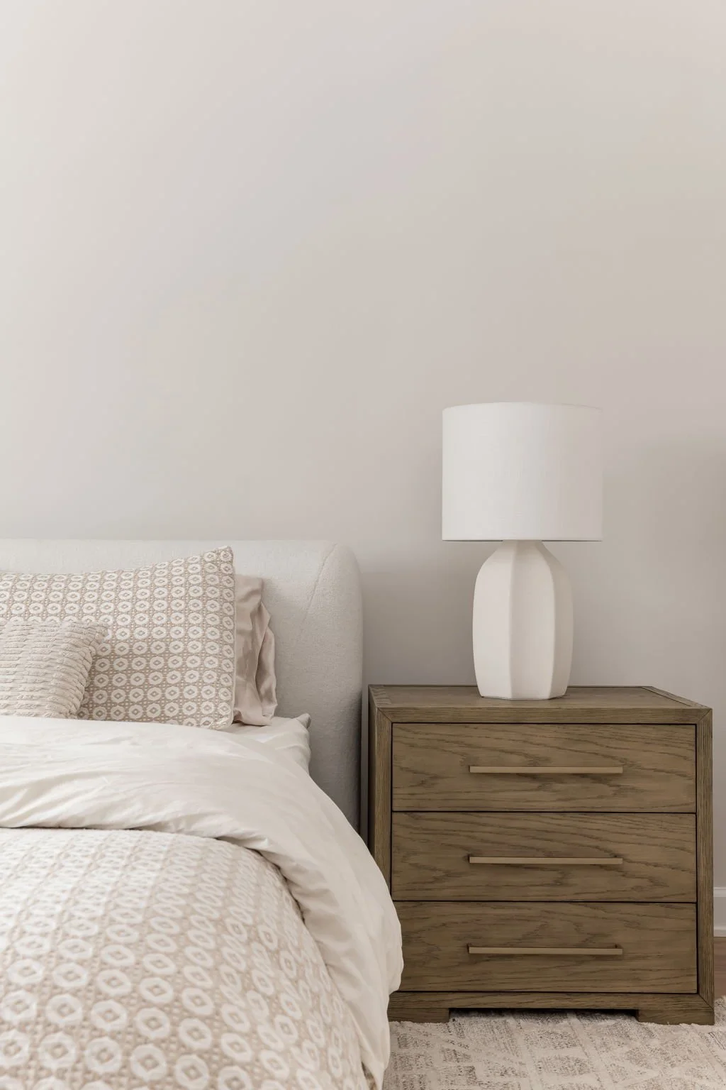 A bedroom with a white upholstered bed, patterned pillows, a wooden nightstand, and a white table lamp on the nightstand, against a plain light-colored wall.