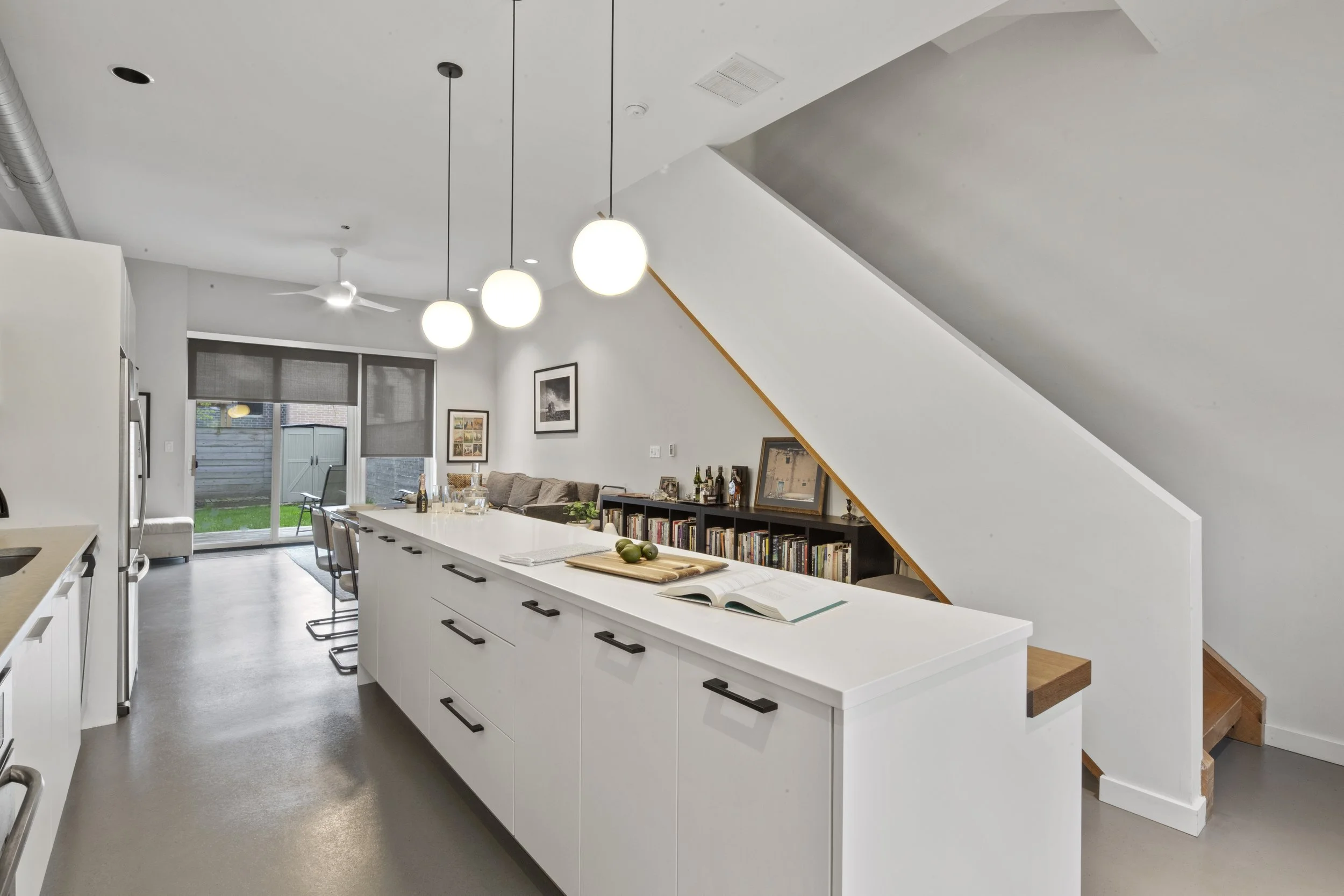 Open-concept kitchen and living room with white cabinets, a large island, pendant lights, a sliding glass door leading to the backyard, and a bookshelf under a staircase.