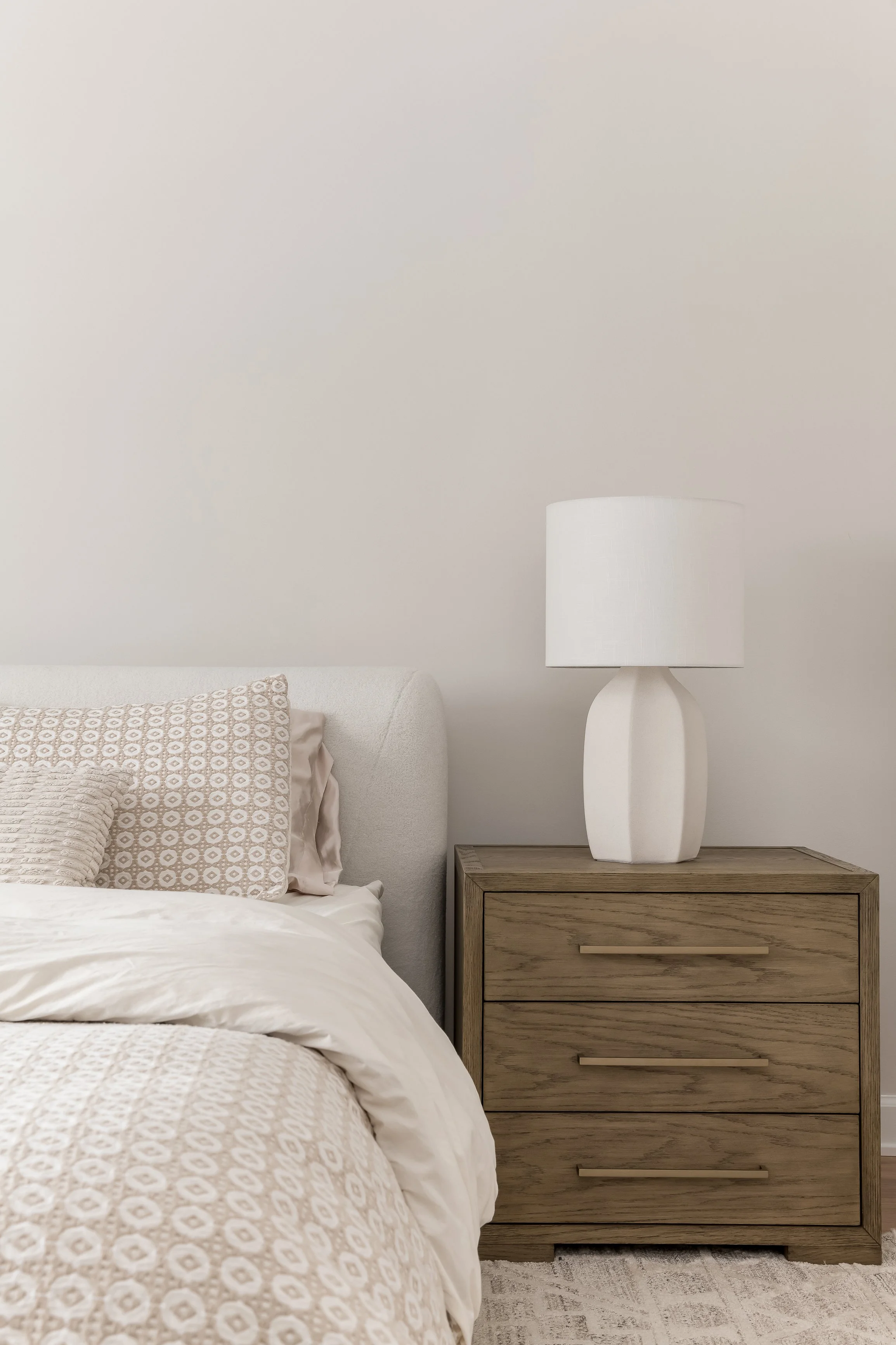 A bedroom with a bed and a wooden nightstand with a white table lamp on it.