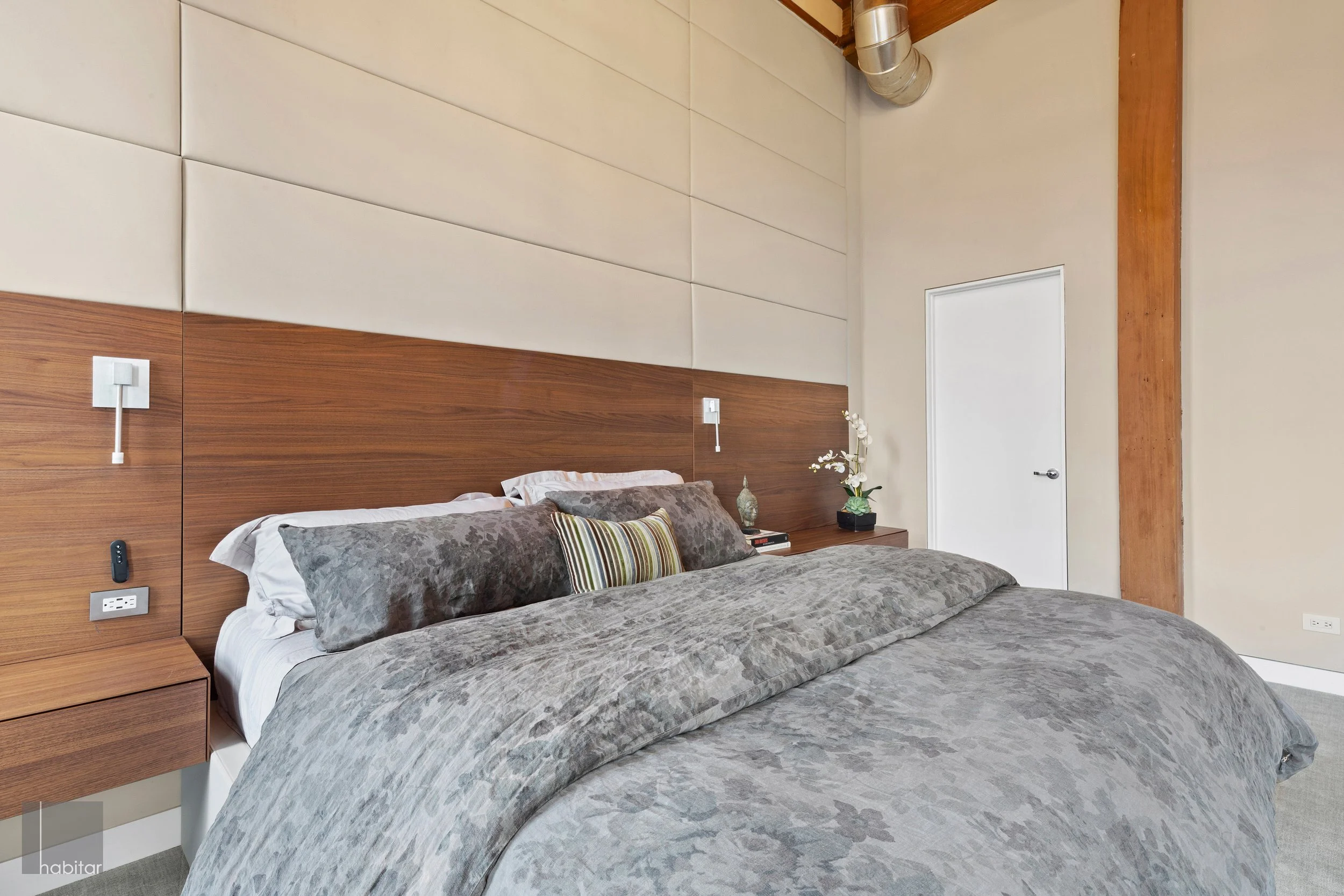 A modern bedroom with a large bed, gray and white bedding, and a wooden headboard. The room features beige walls, a white door, and a window out of view. Nightstands with lamps and decorative items flank the bed.