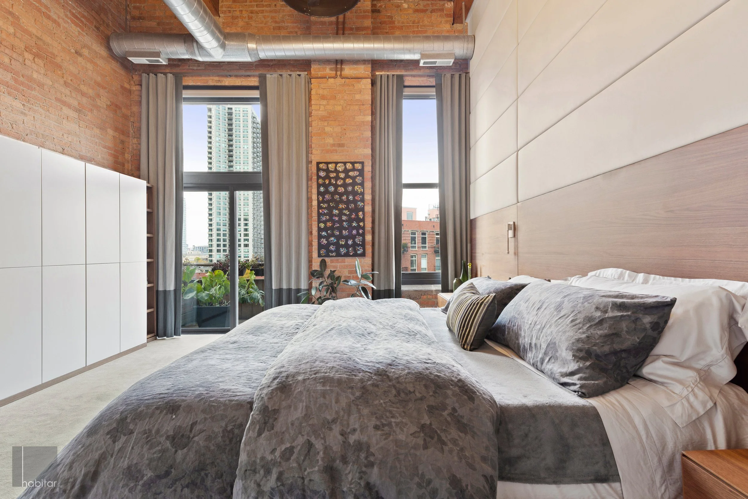 A bedroom with a large bed, decorative pillows, a wooden headboard, and tall windows showing city buildings outside. There are plants on the window sill and a piece of artwork on the brick wall. The room has an industrial style with exposed ductwork.