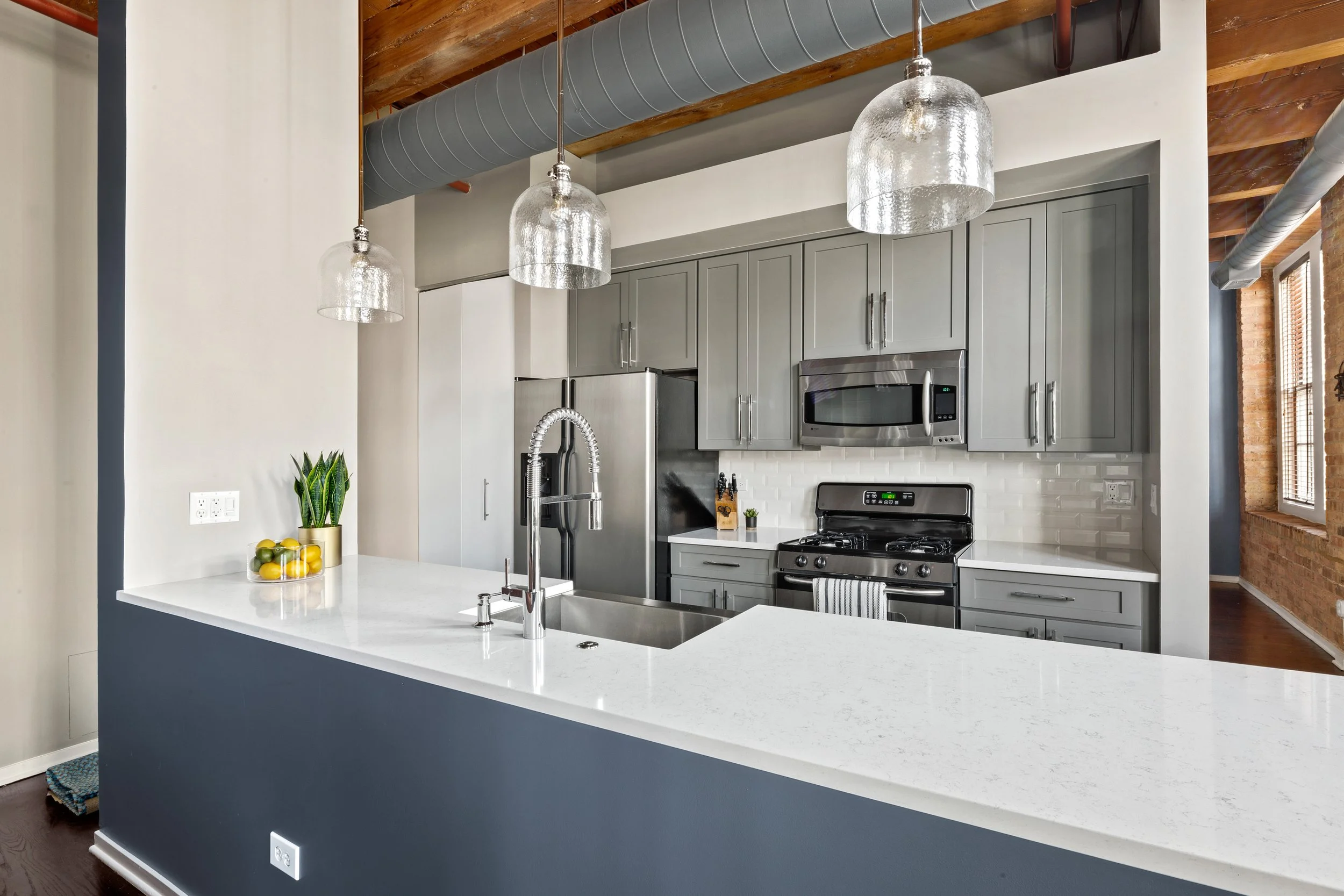 Modern kitchen with gray cabinets, stainless steel appliances, white countertop, green plant, and hanging glass pendant lights.