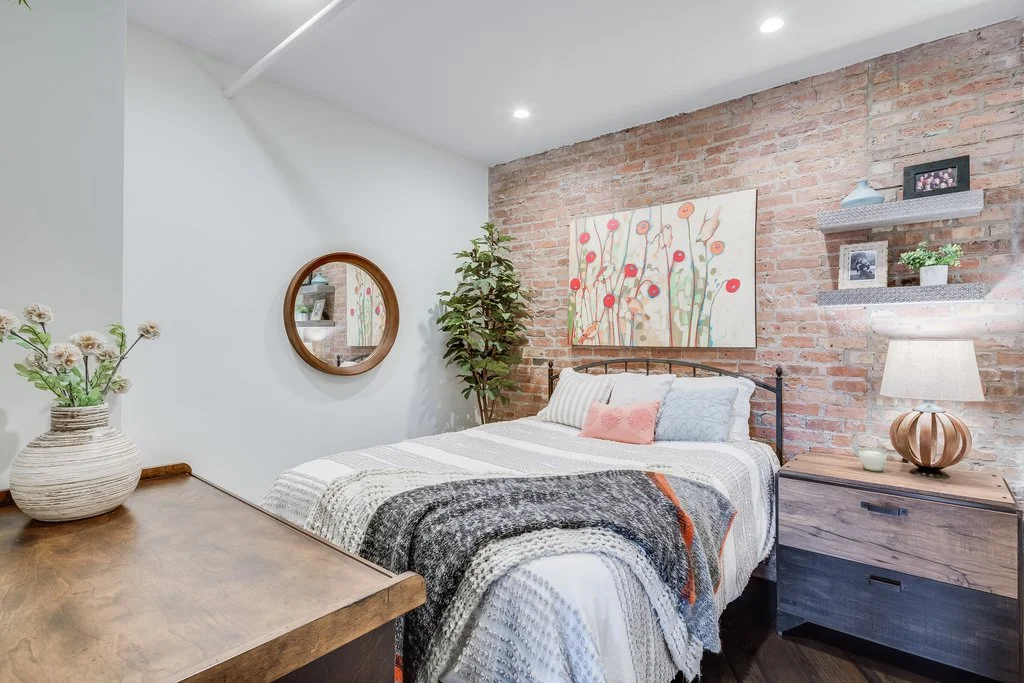 Cozy bedroom with a bed covered in a white and gray blanket, pink and gray pillows, and a floral painting over a brick accent wall. A nightstand with a lamp and decorative items, a large potted plant, and a round mirror hanging on a white wall are al