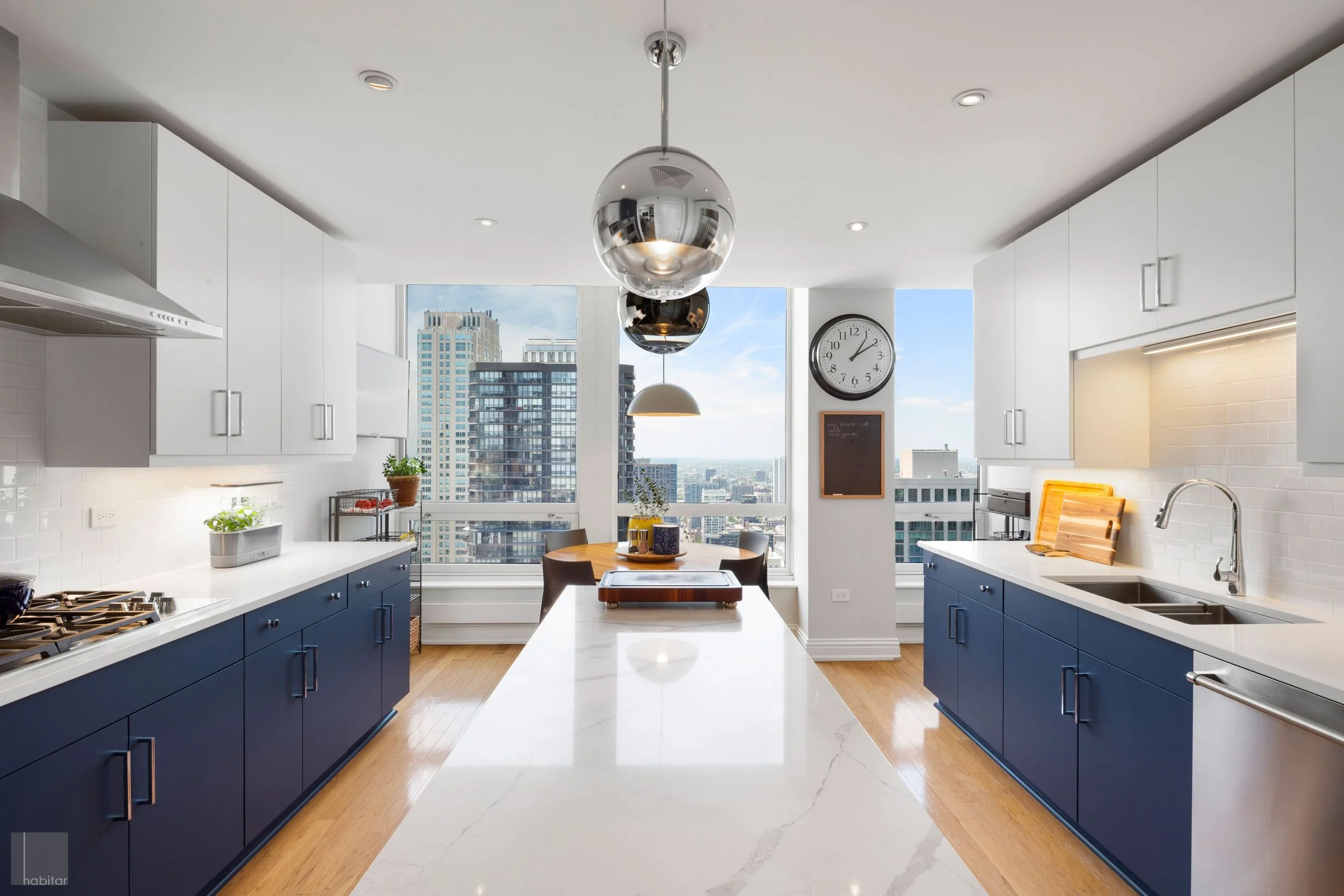 Modern kitchen with white upper cabinets, dark blue lower cabinets, and a large marble island in the center, overlooking a cityscape through large windows.