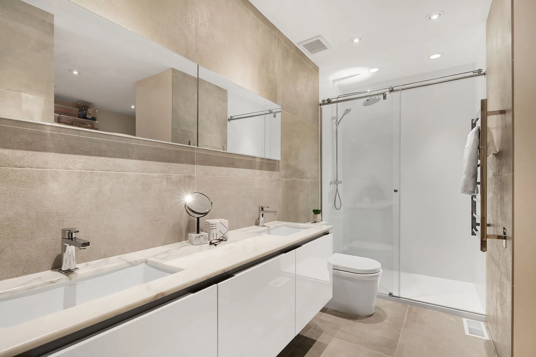 Modern bathroom with double sink vanity, large mirror, walk-in shower with glass door, toilet, and beige tiled walls and floor.