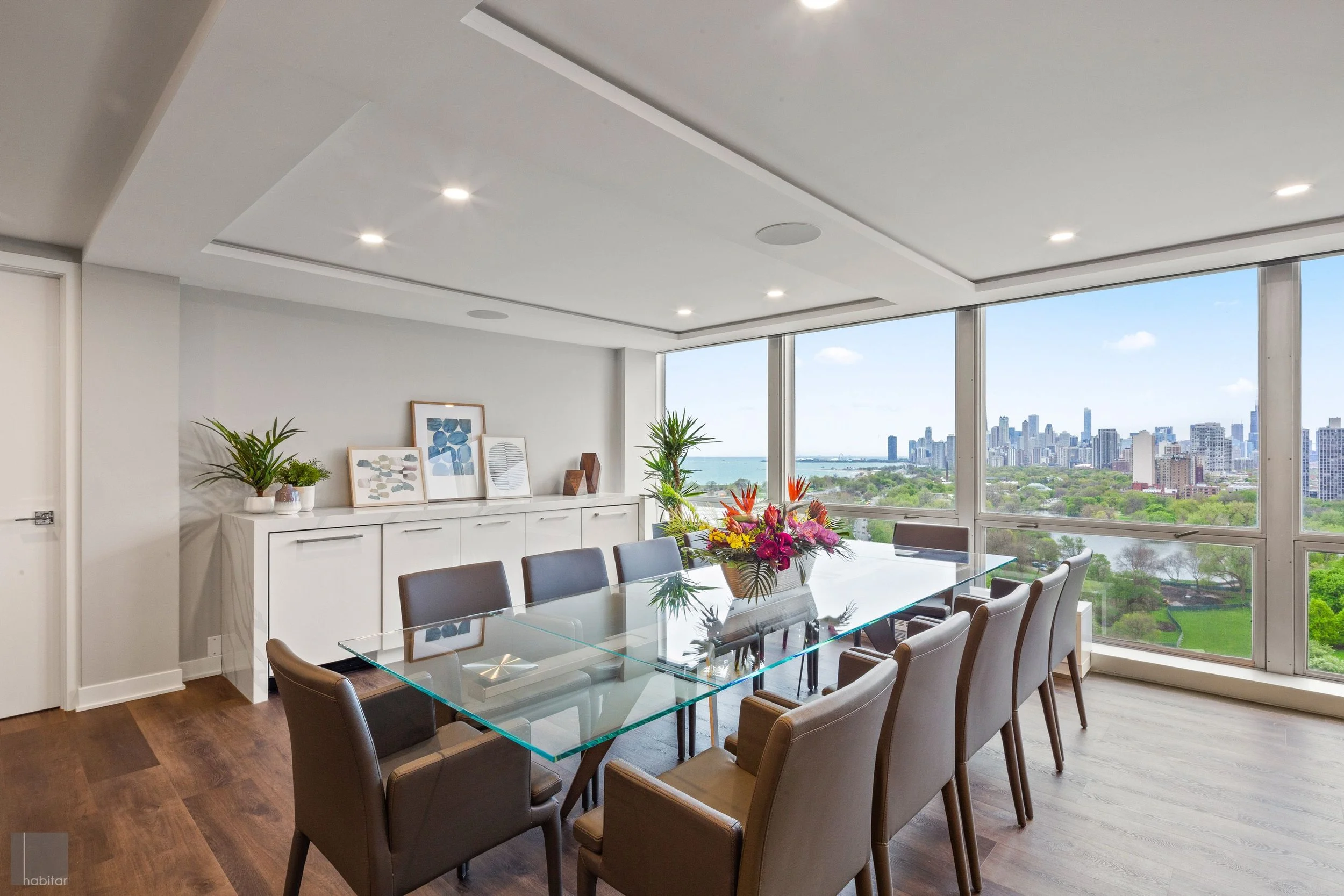 Modern high-rise dining room with glass table, 10 chairs, and a view of the city skyline through large windows, decorated with plants and artwork.