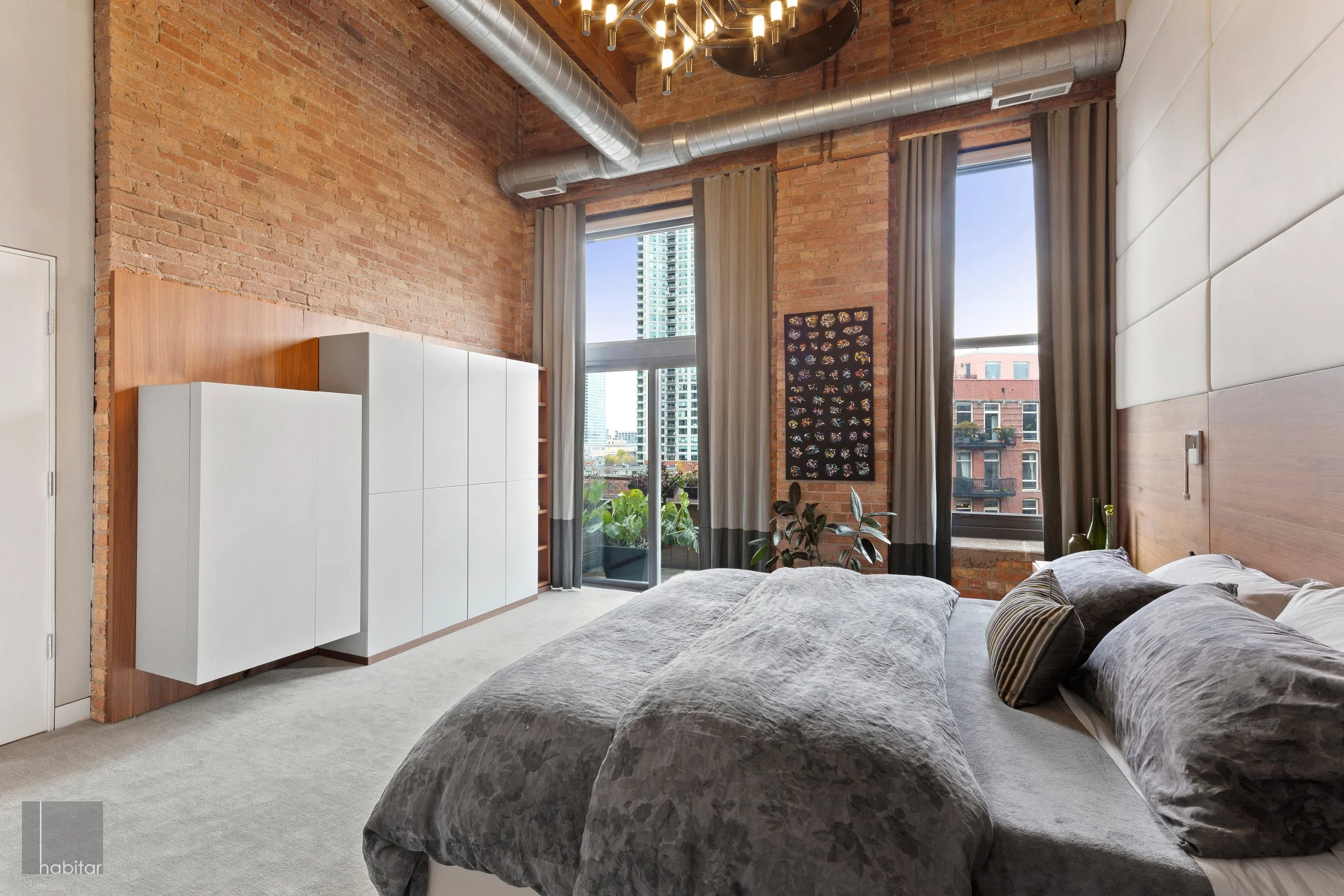 Modern bedroom with exposed brick walls, large windows with gray curtains, a bed with gray bedding, and a city view outside.