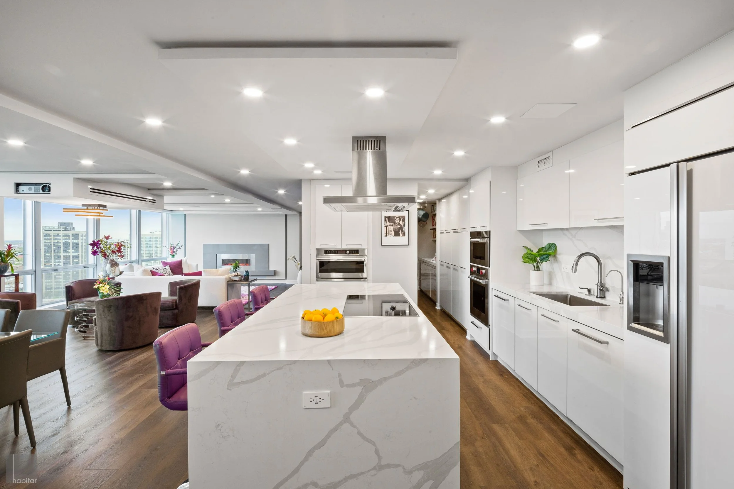 Modern open-concept kitchen and living area with white cabinetry, marble countertops, and purple seating, overlooking city skyline.