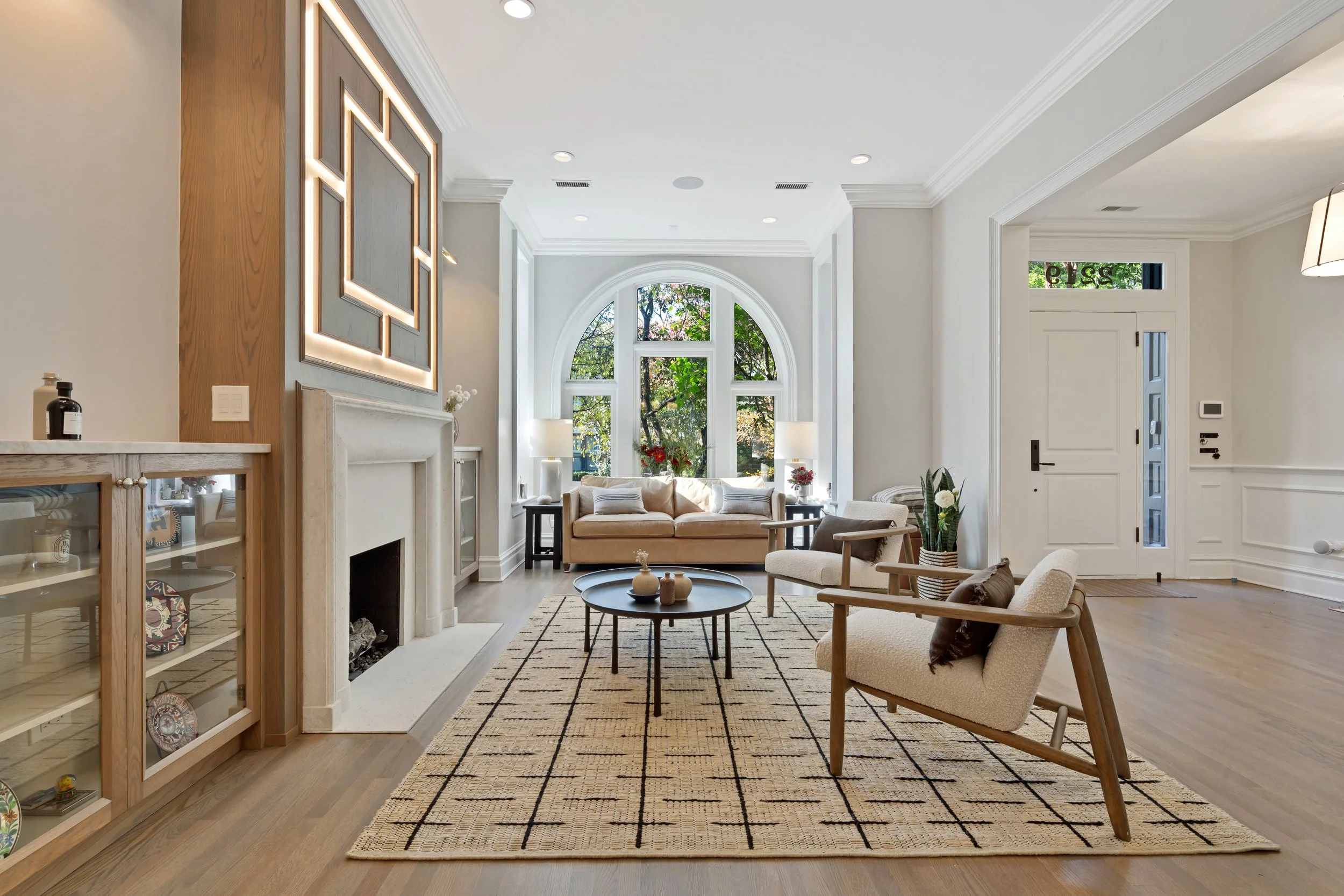 Bright living room with large arched window, beige sofa, and armchairs, wooden floor, rug, fireplace, and modern decor.